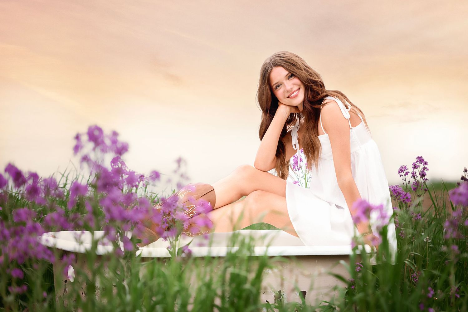 Woman in white dress poses gracefully in a field of vibrant purple wildflowers during golden sunset.
