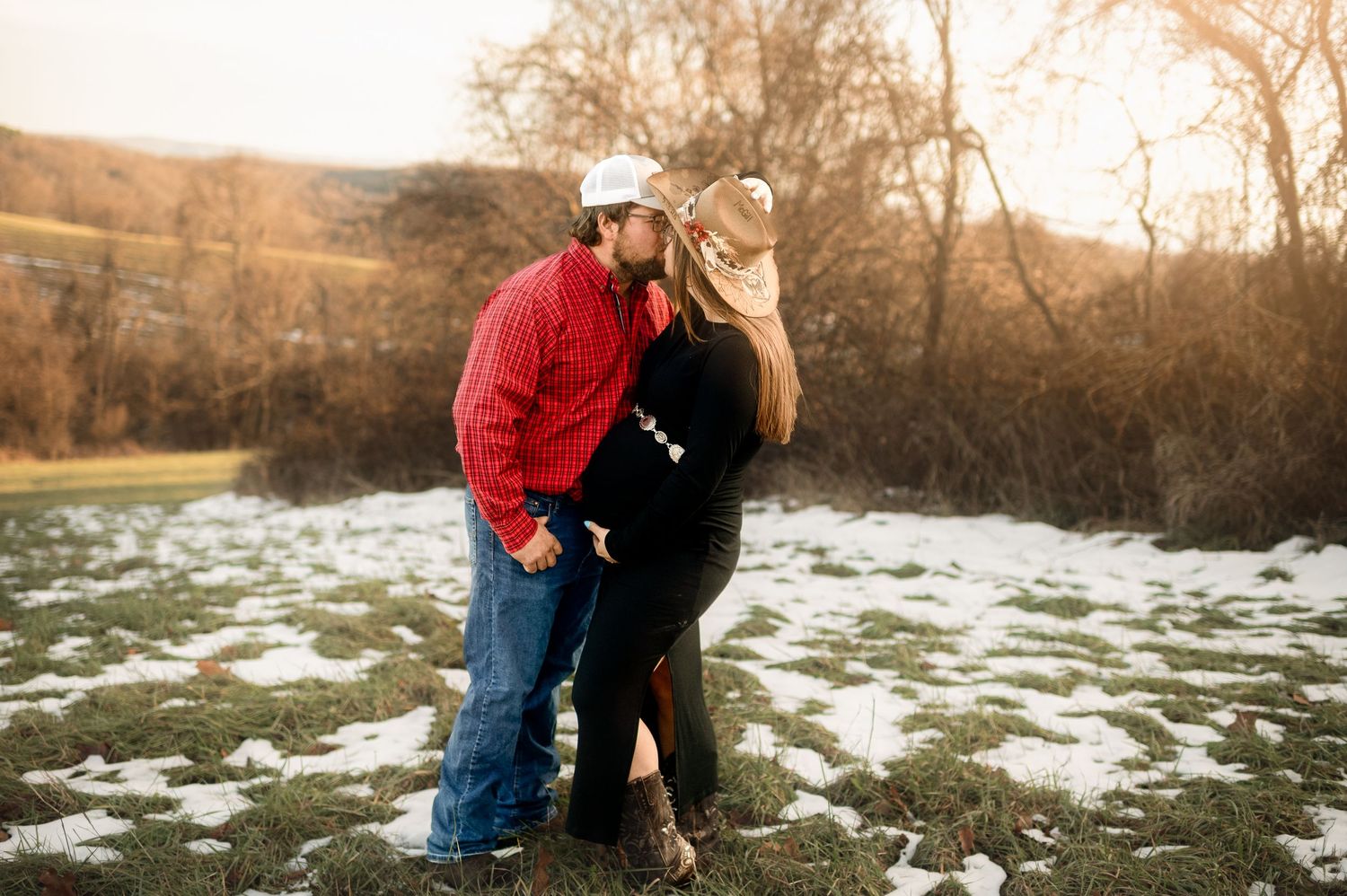 A couple embraces in a snowy winter landscape at sunset with bare trees and hills in the background.
