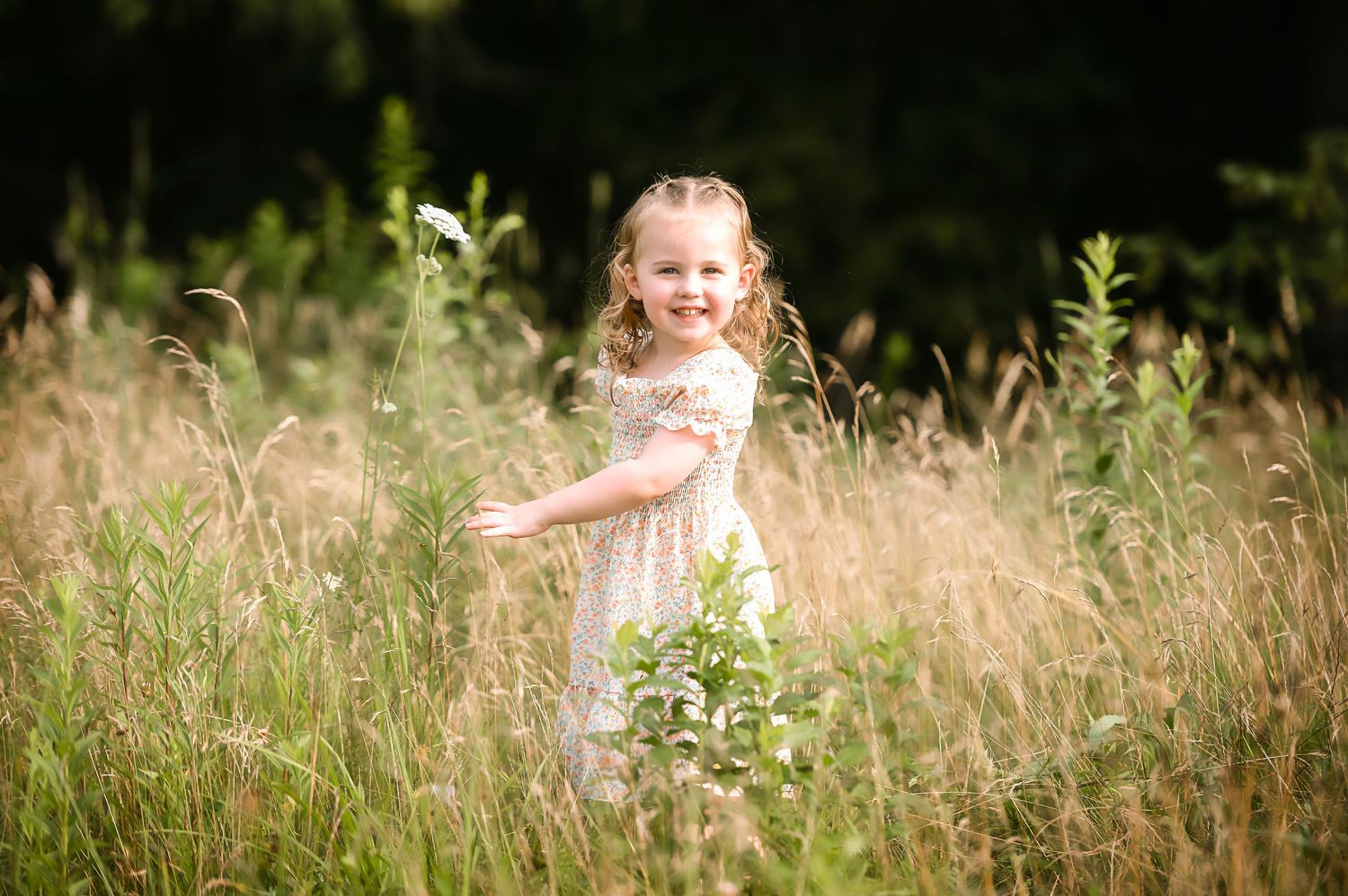 A child in a white dress sits joyfully in a sunlit meadow surrounded by tall grass and wildflowers.