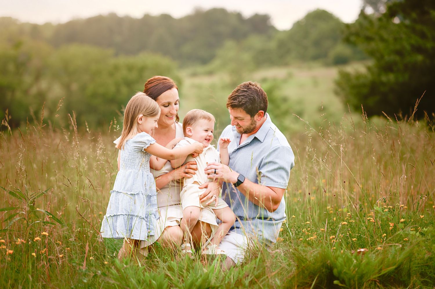 Family sharing tender moments together while sitting in a sunlit meadow with tall grass and wildflowers.