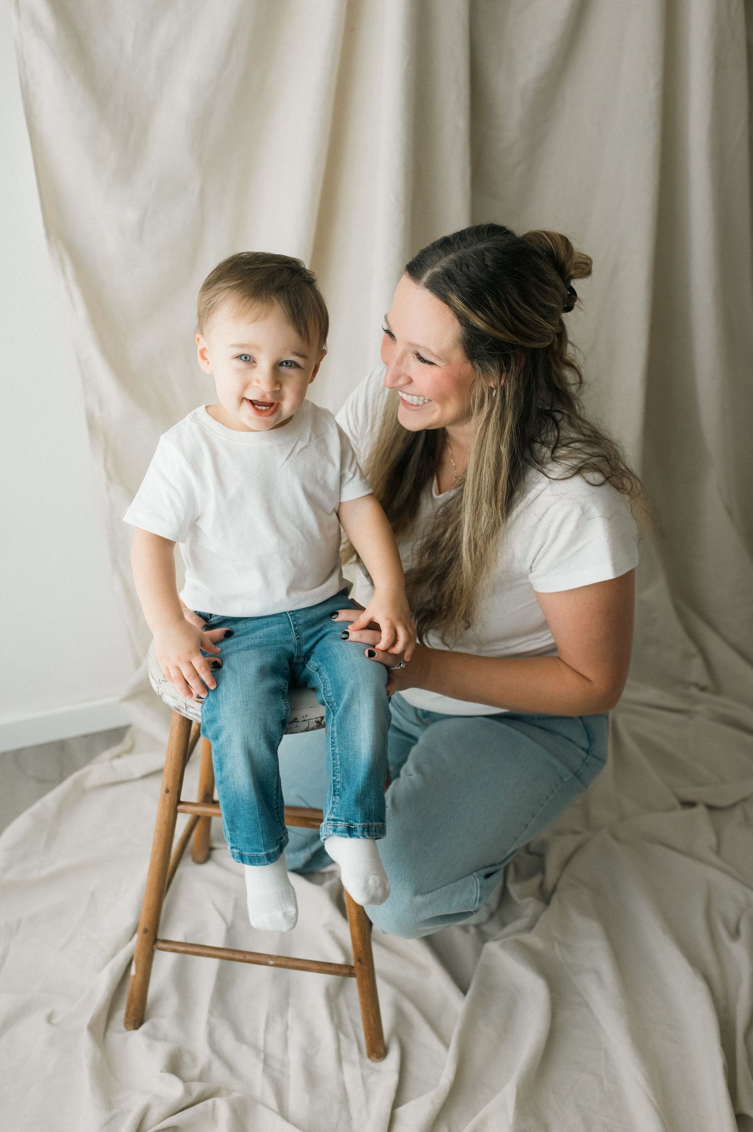 A tender moment captured against a white backdrop as they share smiles while sitting on a wooden stool.