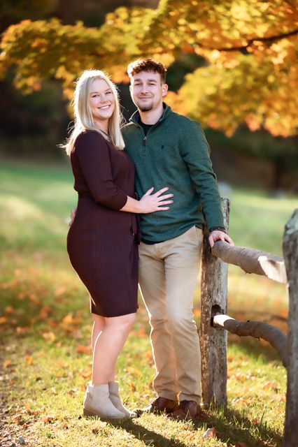 A couple stands together by a wooden fence surrounded by golden fall leaves.