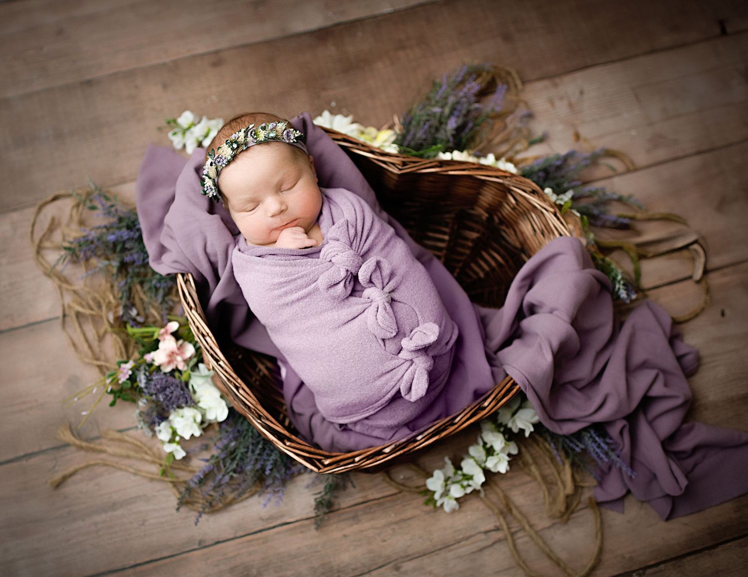 Sleeping newborn wrapped in purple fabric lies in wicker basket surrounded by delicate flowers and greenery.