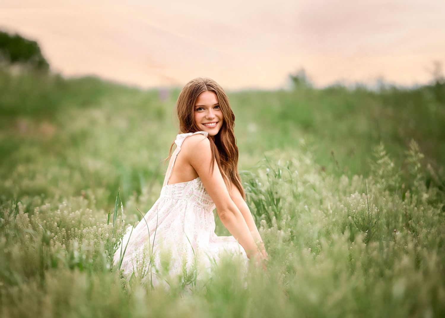 A person in a flowing white dress sits gracefully in a sunlit meadow of tall grass and wildflowers.