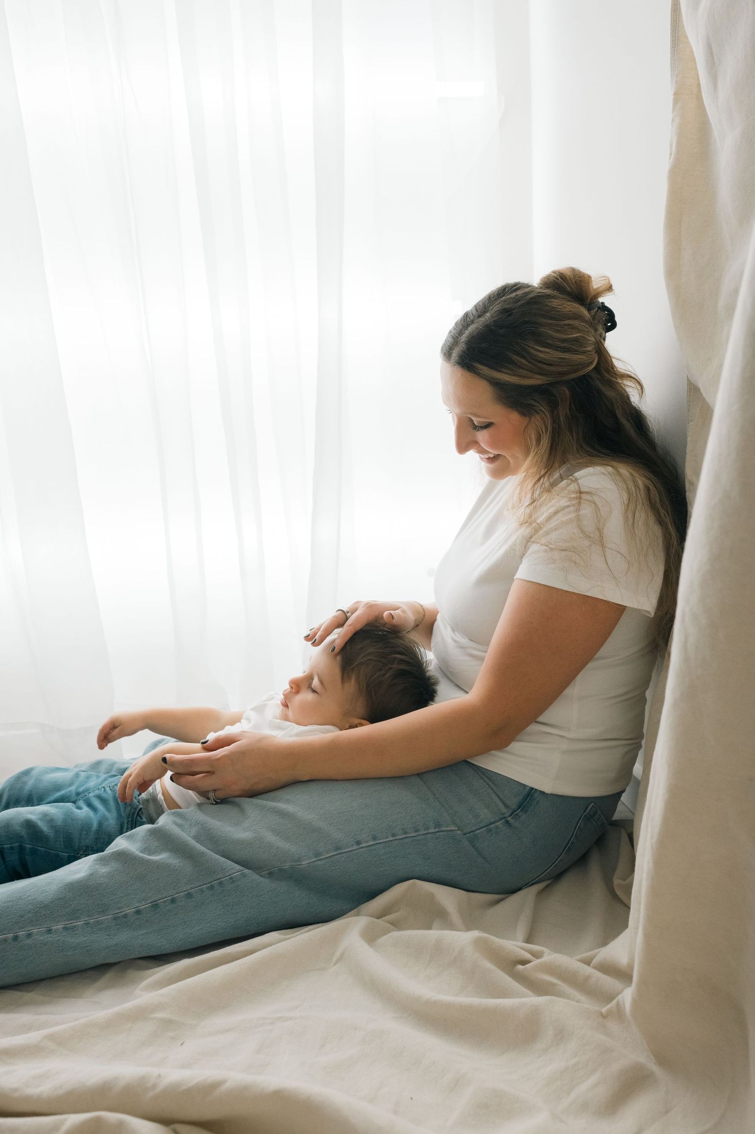 Parent and child share tender moment while relaxing together on white bedding in natural light.