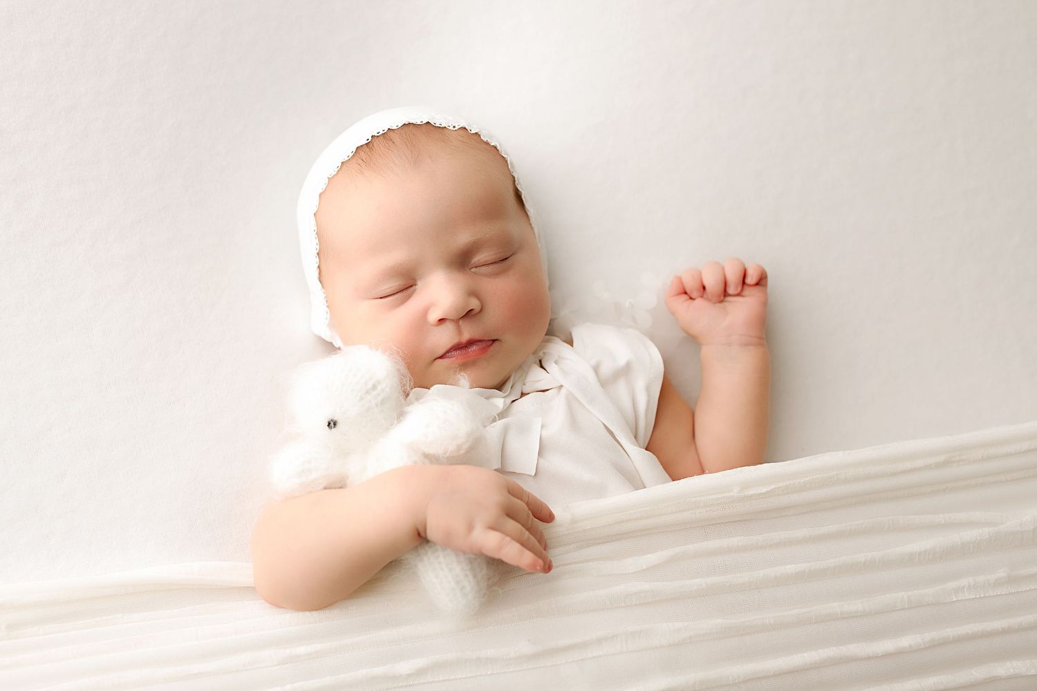 A sleeping baby in white clothing rests peacefully on white bedding in a bright, minimalist setting.