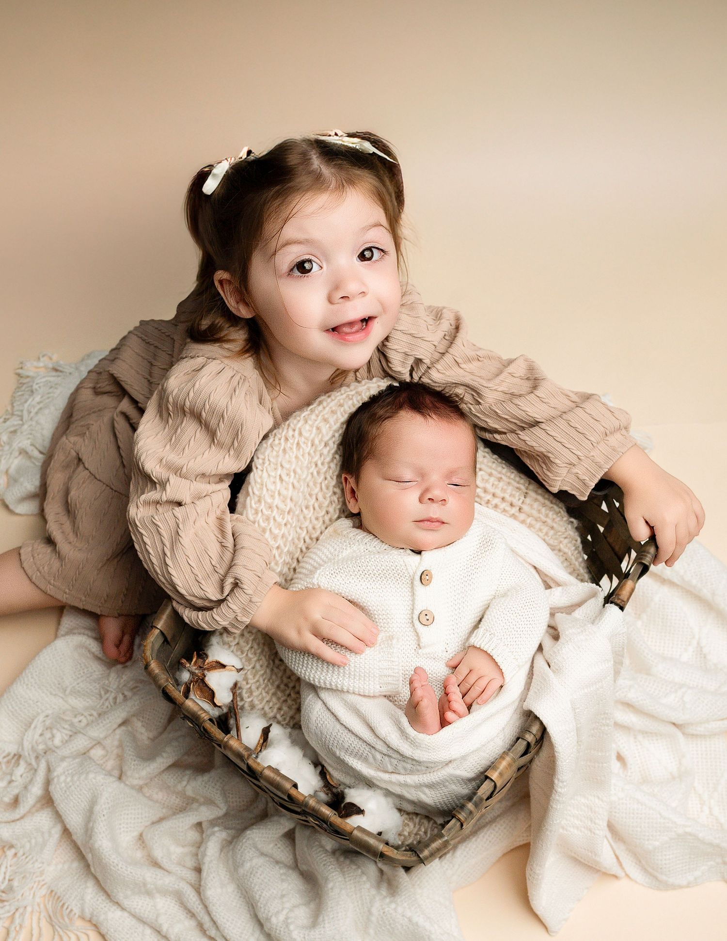 Sibling portrait with newborn baby in cream knit outfit on textured neutral backdrop.