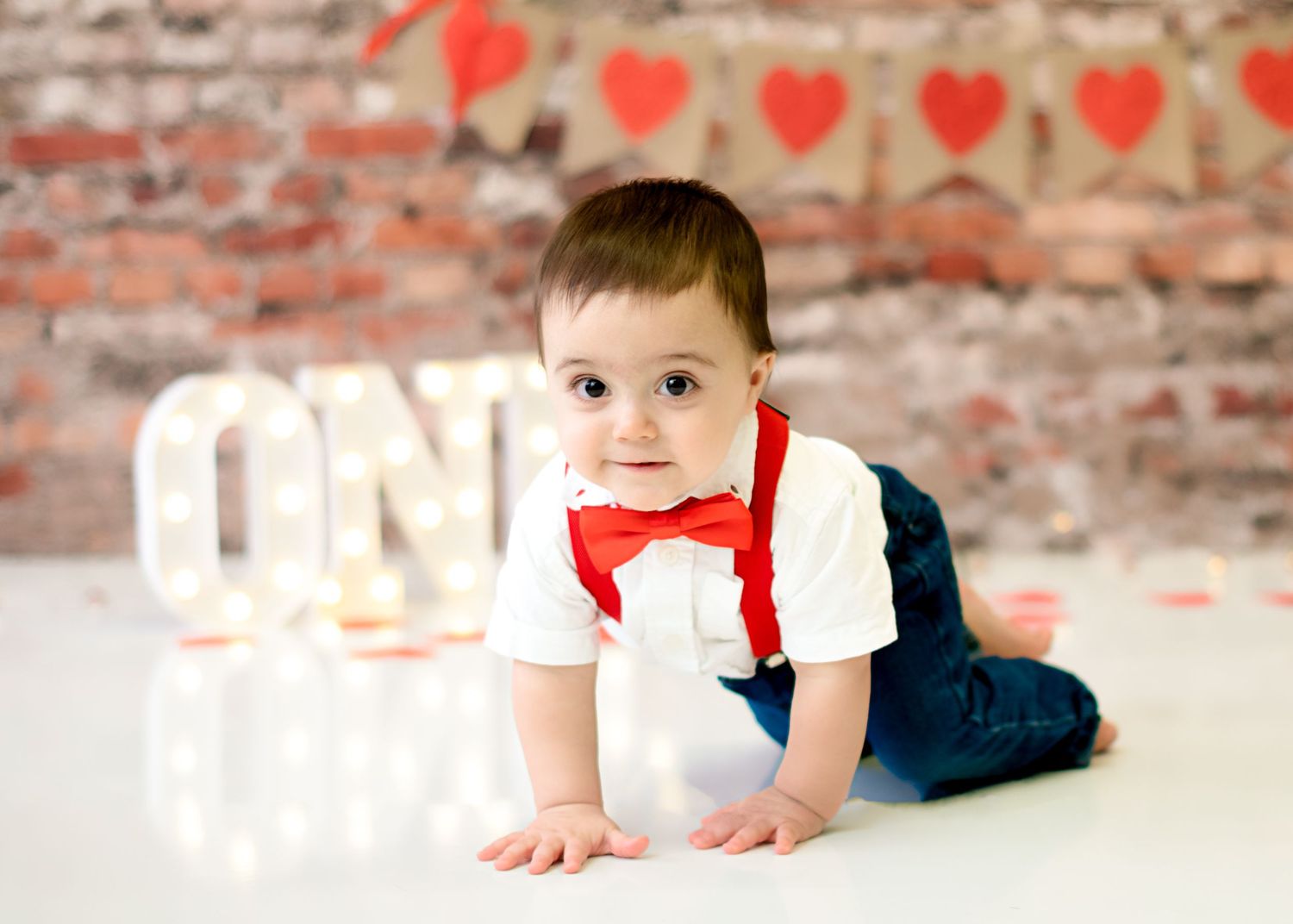 Baby in formal outfit with red bowtie crawling on floor with heart decorations in background.