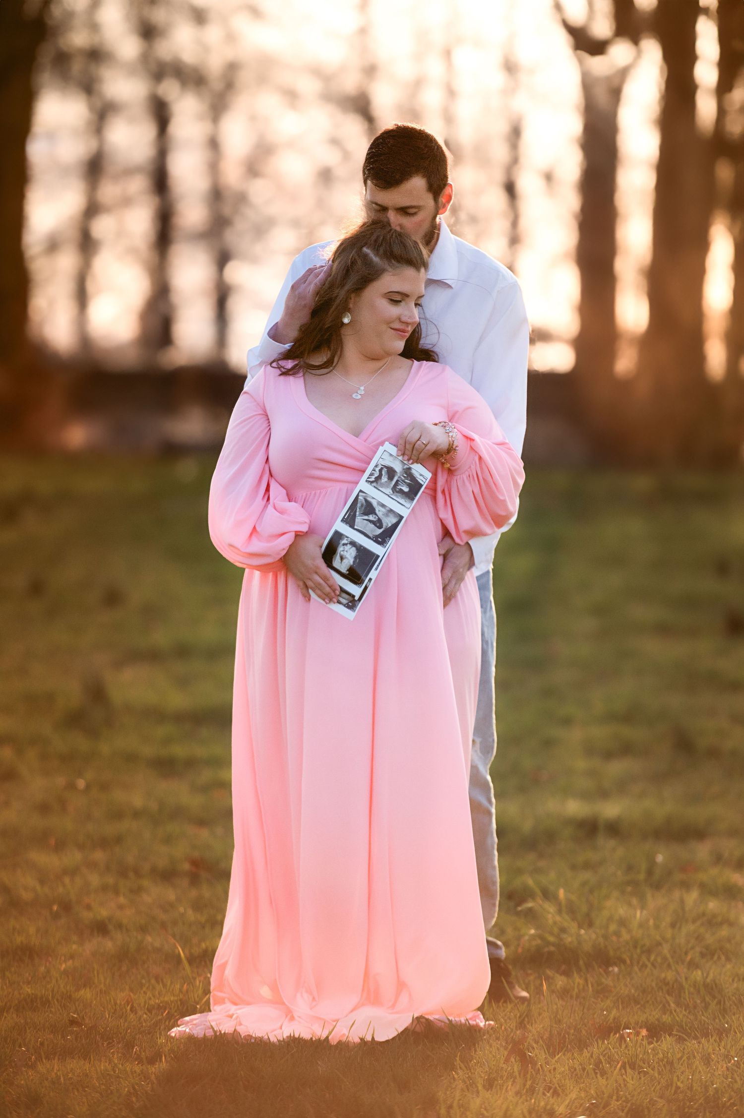 A maternity photoshoot in a park at sunset with a woman in a pink flowing dress.