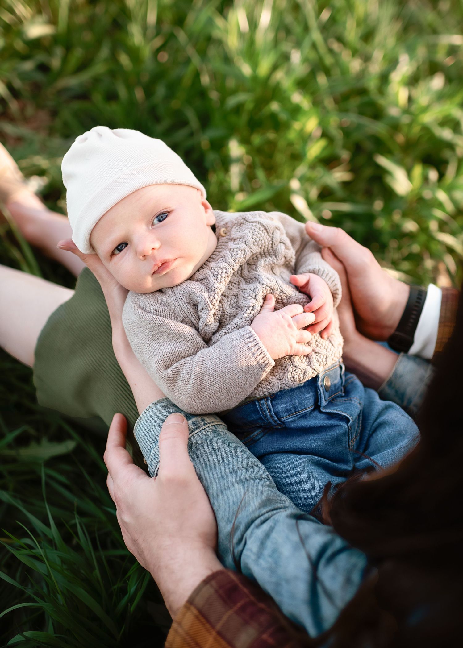 Baby in sweater and jeans held by hands against grass background.