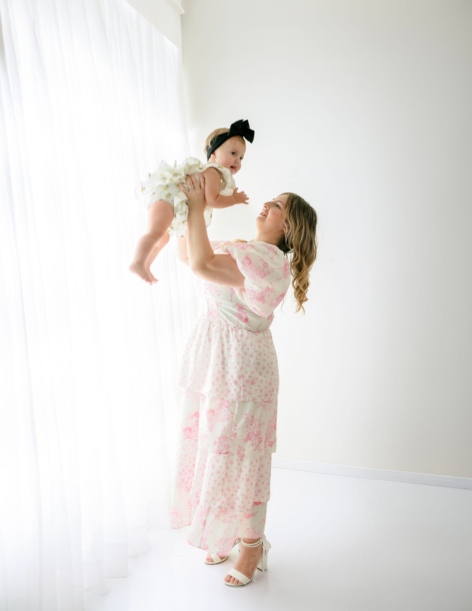 Mother in pink floral dress lifts baby wearing white outfit against bright white wall.