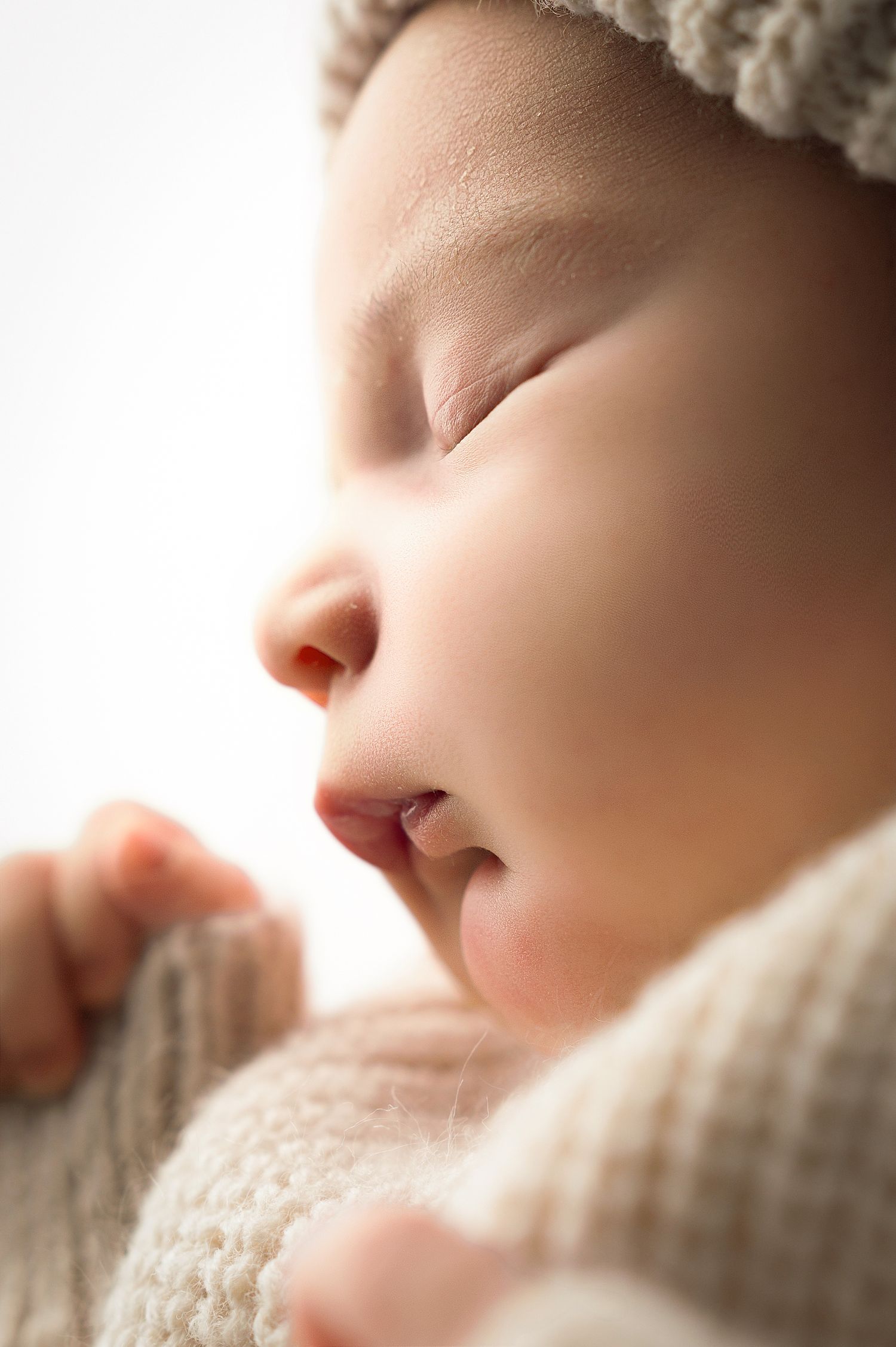 Close-up profile view of a sleeping baby wearing a soft white knit hat against a light background.