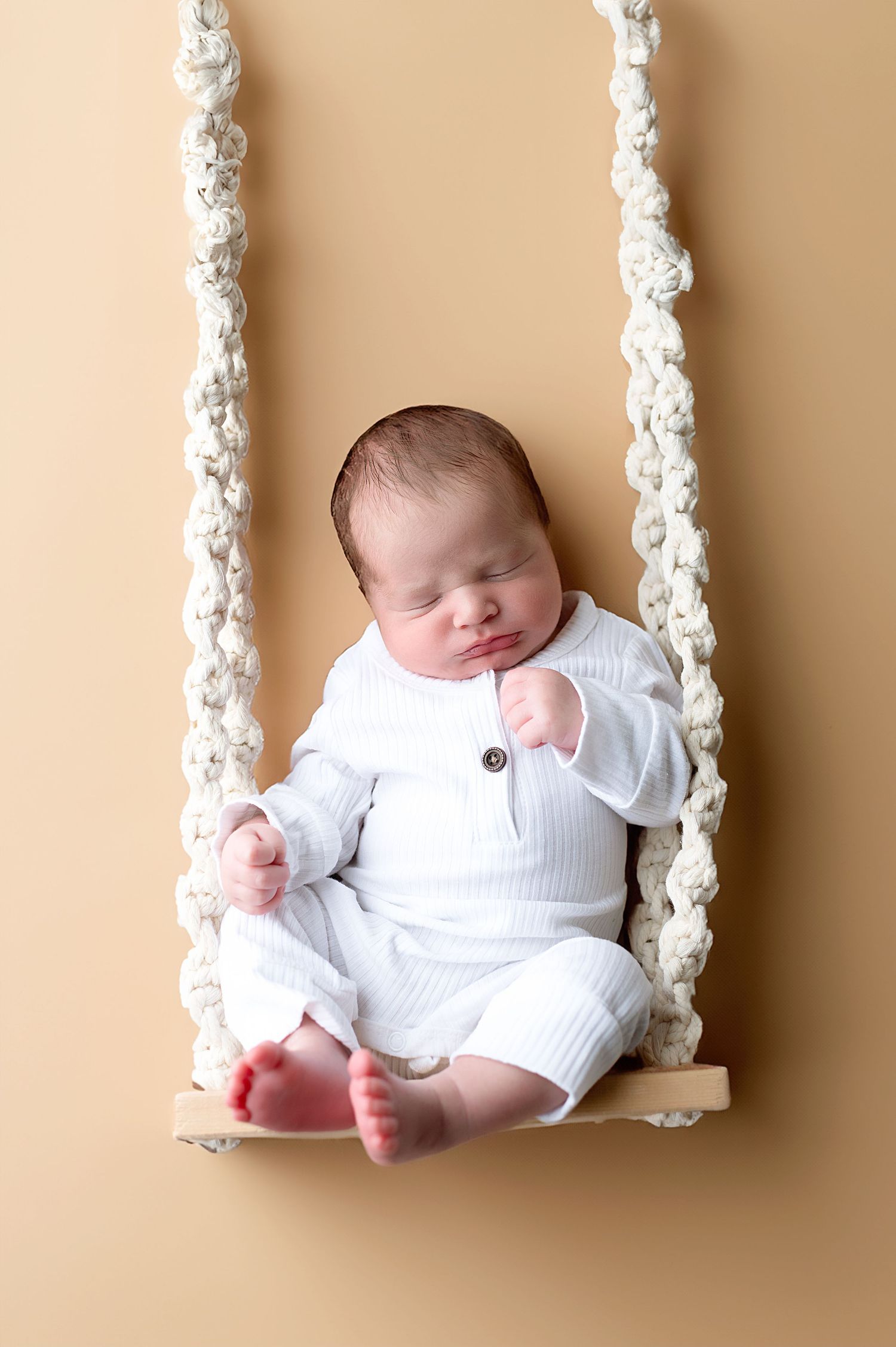 Baby in white outfit sleeping peacefully on macrame rope swing against beige background.