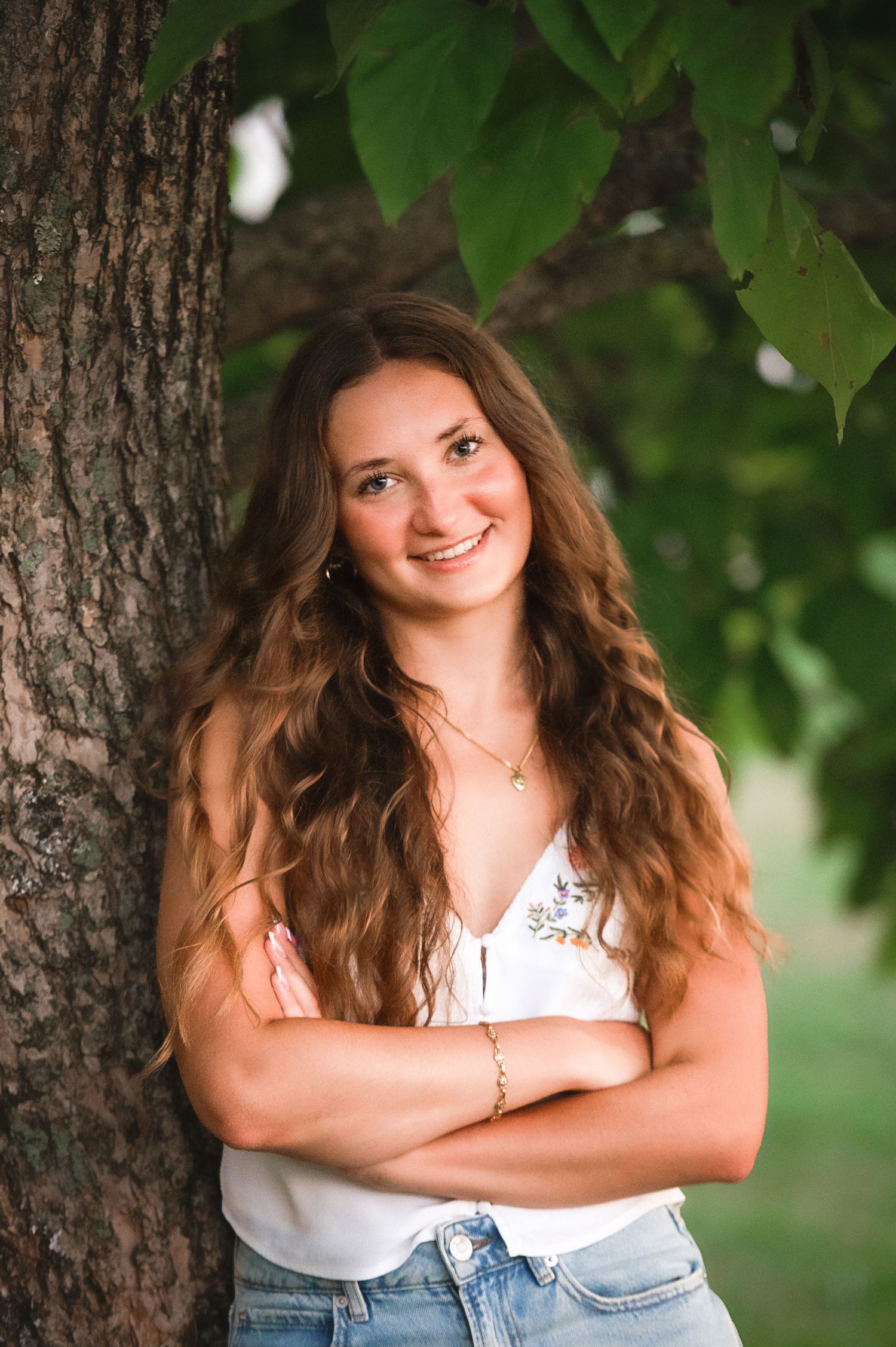 Portrait of someone in casual attire leaning against a tree trunk with green leaves in the background.