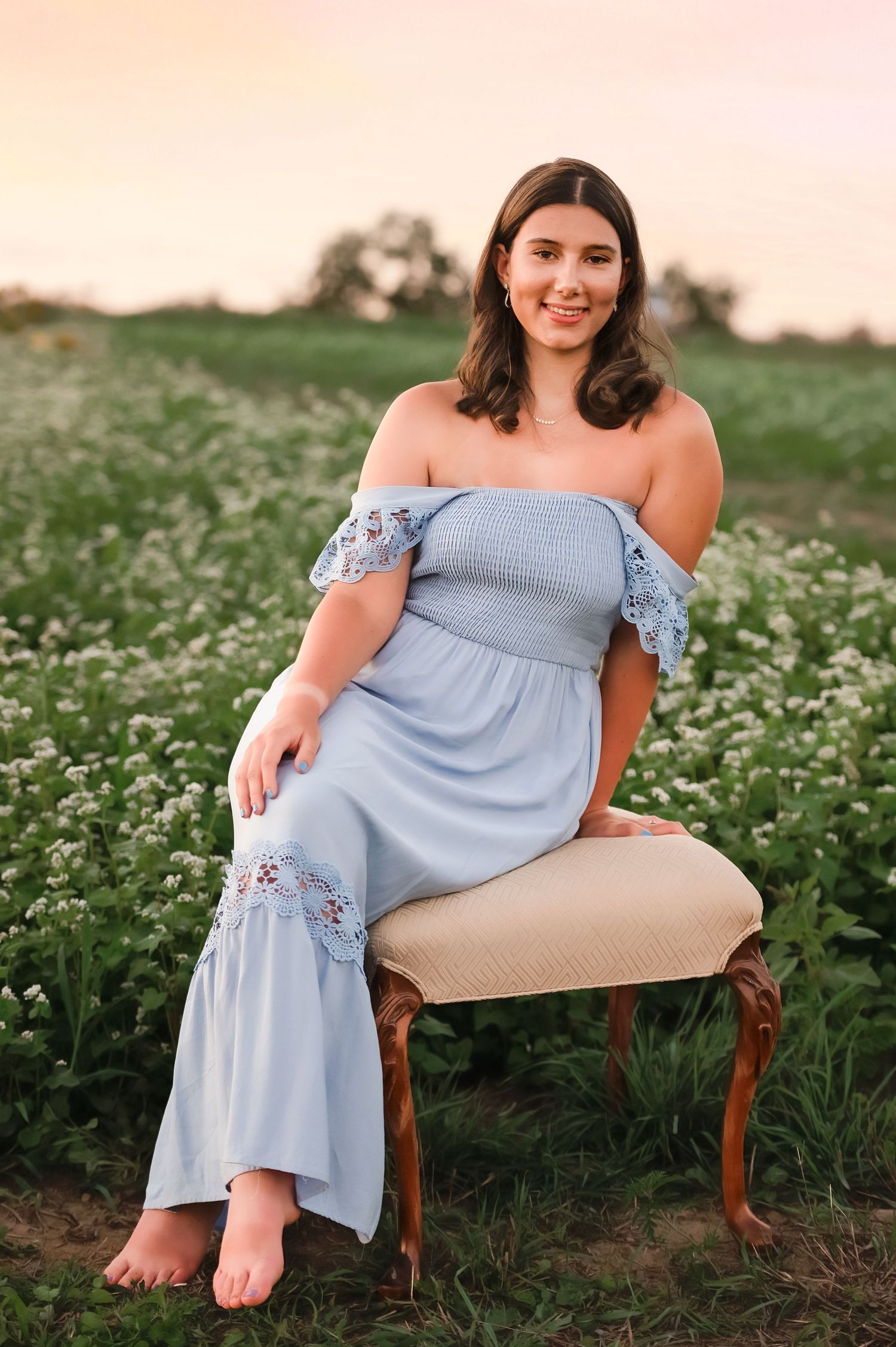 Person in a light blue off-shoulder dress sitting on an antique chair in a flower field.