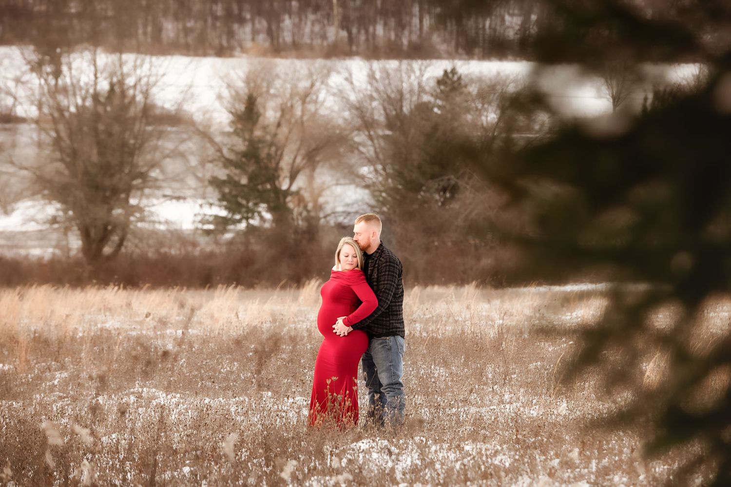 A couple poses together in a snowy winter field with tall dried grass and bare trees in the background.
