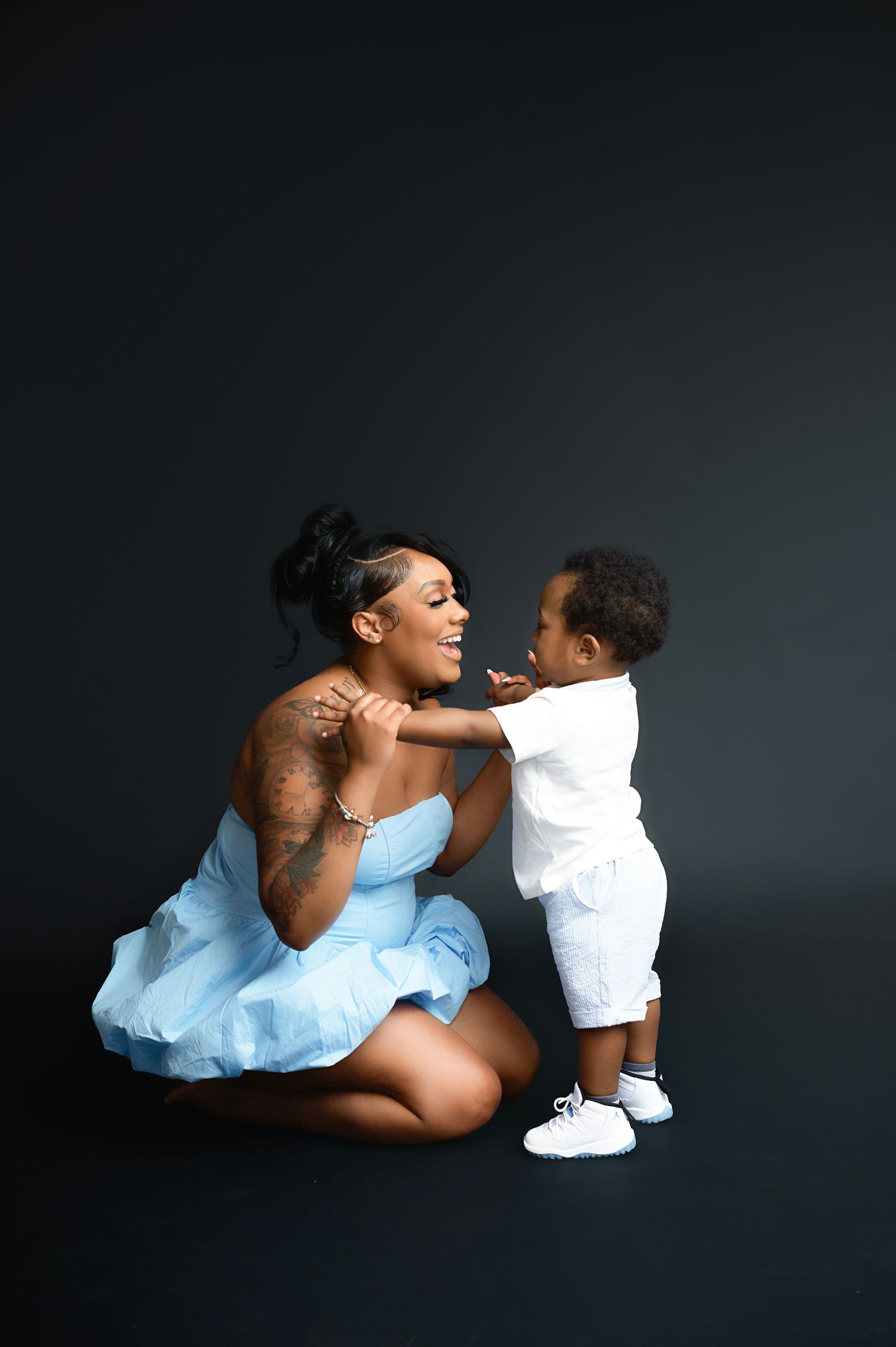 Playful interaction between parent in blue dress and child in white outfit against dark backdrop.