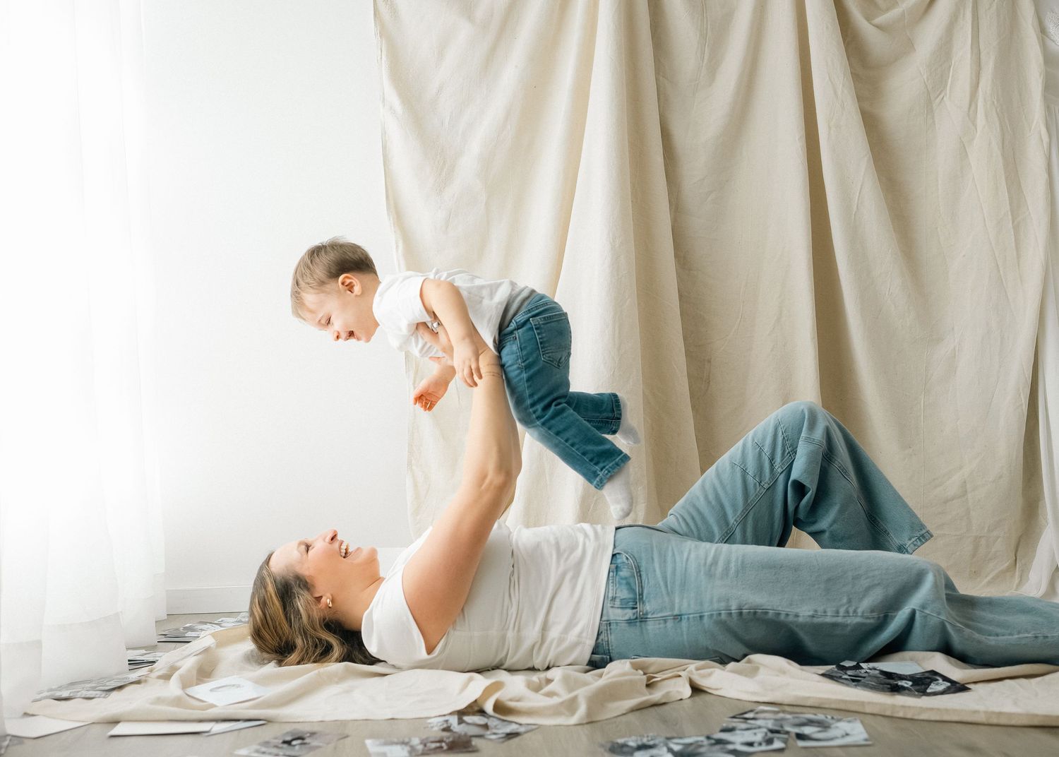A person in blue jeans lies on a white bed while reading a book in a bright, airy room.