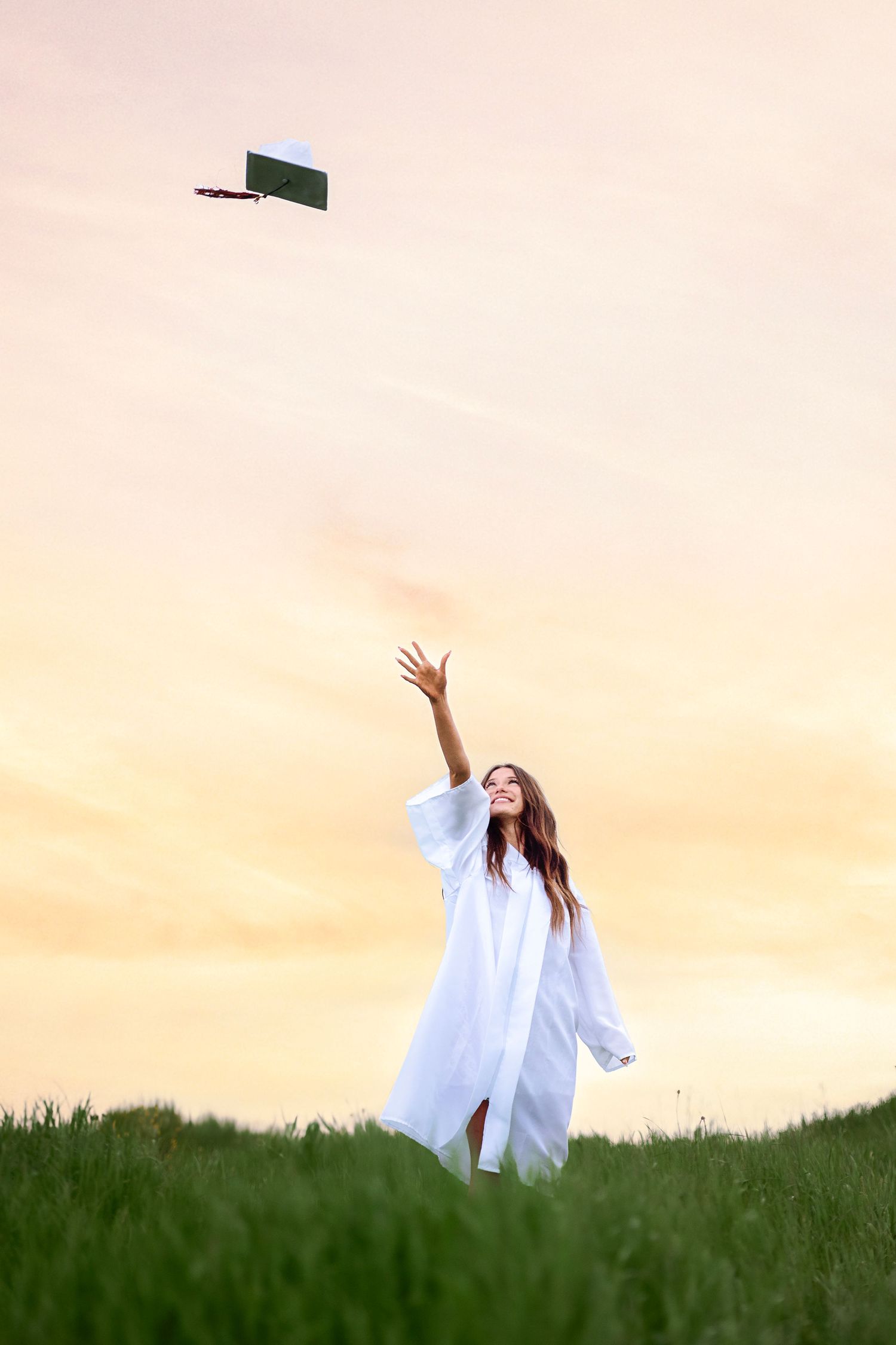 Graduate in white gown tosses cap into sunset sky on green hill, celebrating academic achievement.