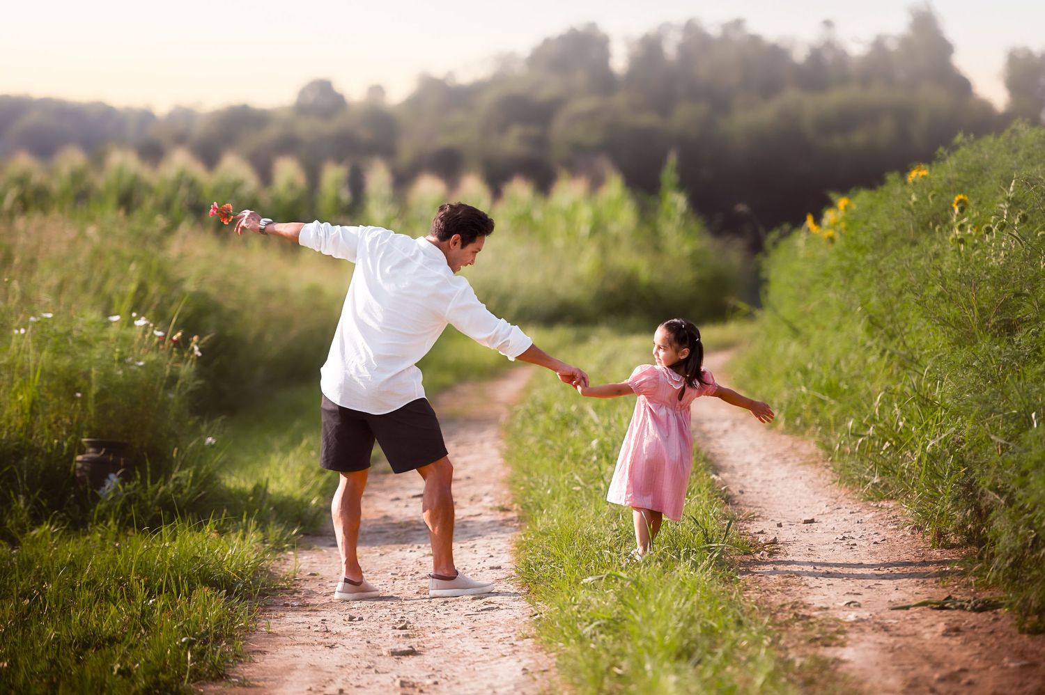 Two people walk playfully down a dirt path between green fields on a summer afternoon.