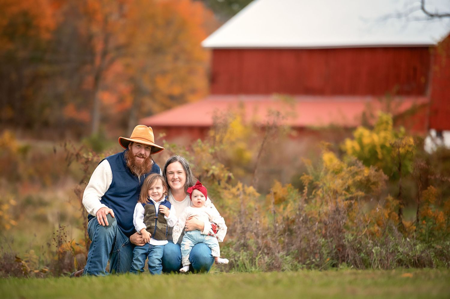 A rustic fall scene with people sitting together near a red barn surrounded by autumn foliage.