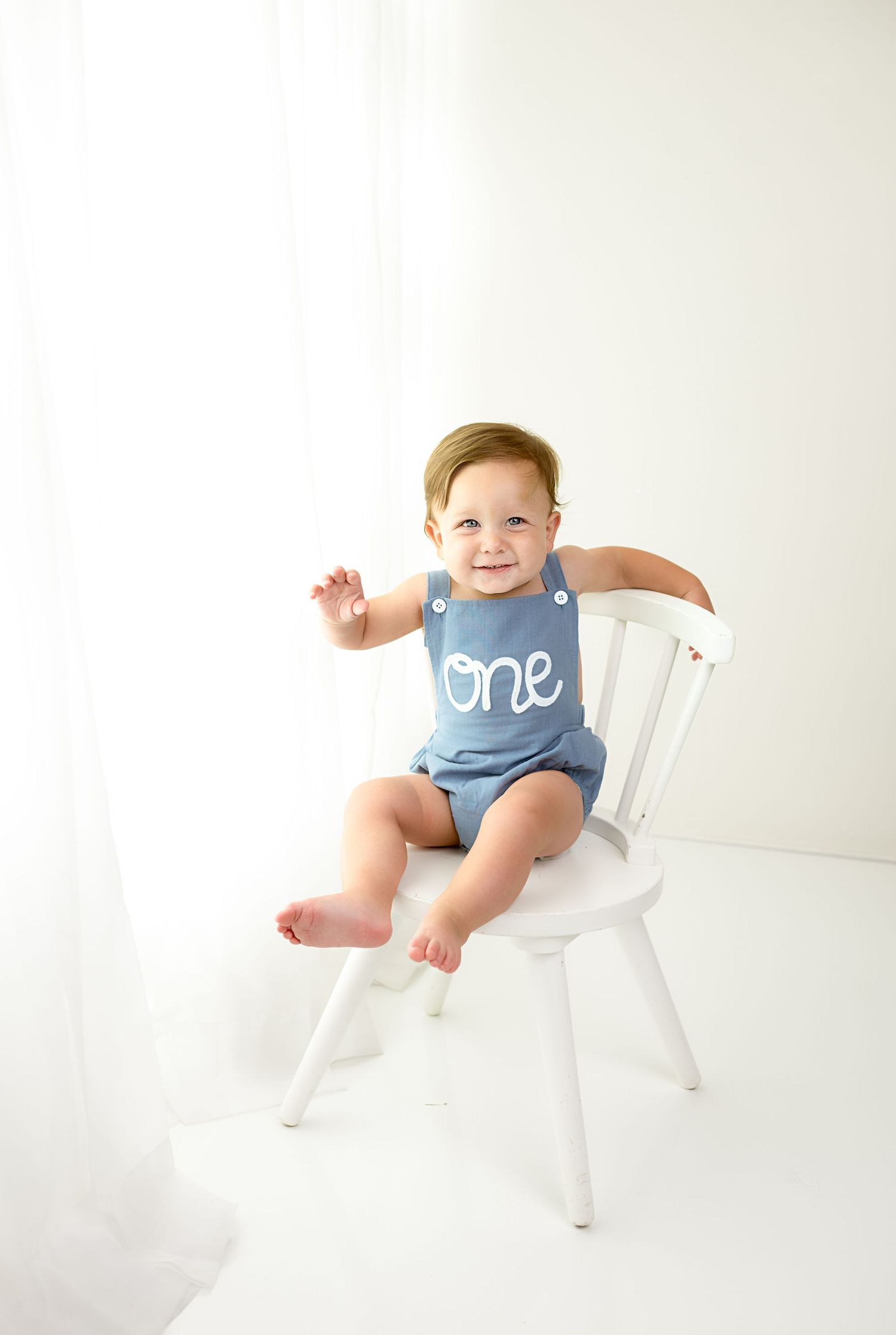 A small child sits in a white wooden chair wearing a blue romper against a white background.