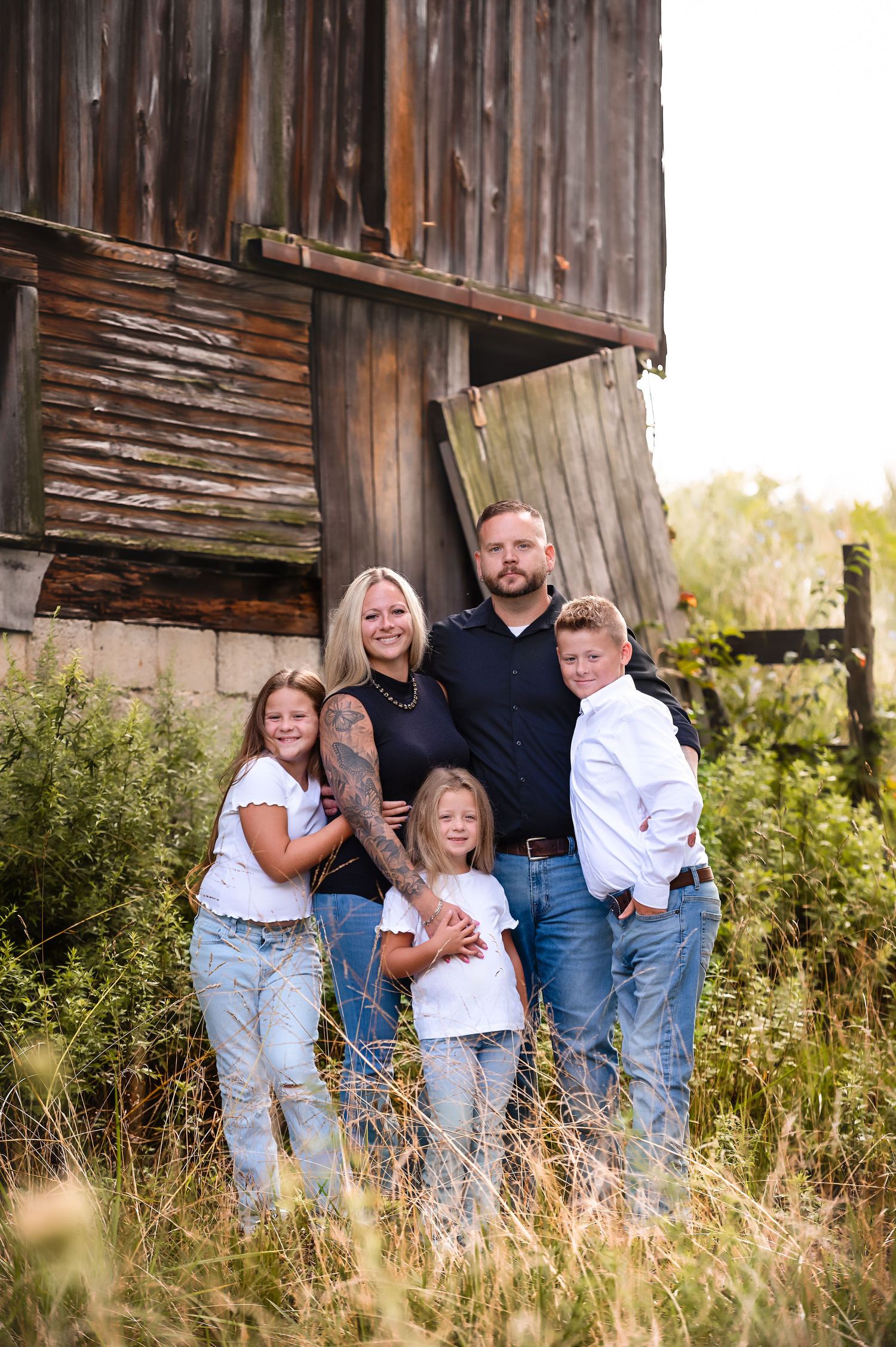 Family portrait session by weathered wooden barn in tall summer grass.