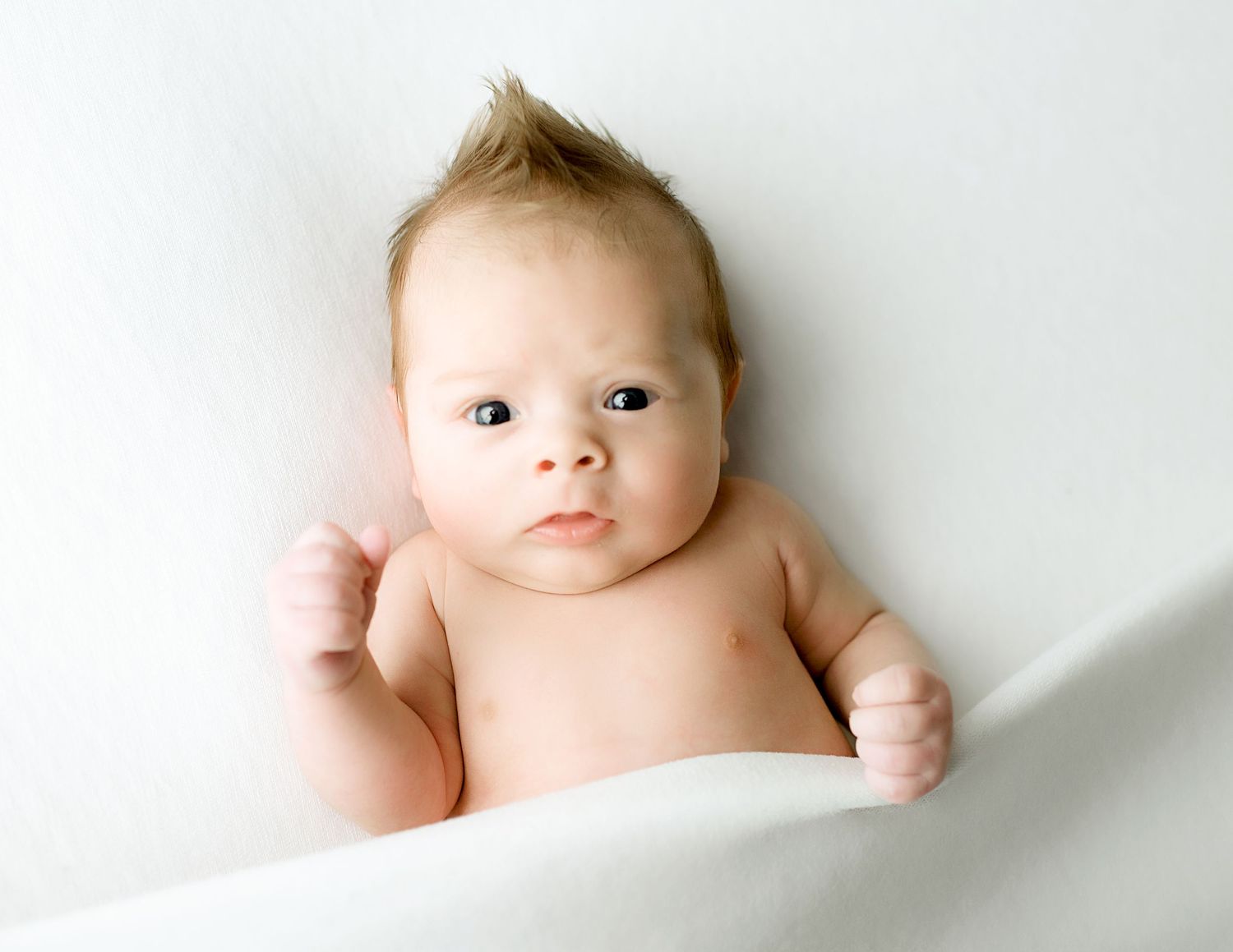 A baby lying on white bedding looking up at the camera with a curious expression.