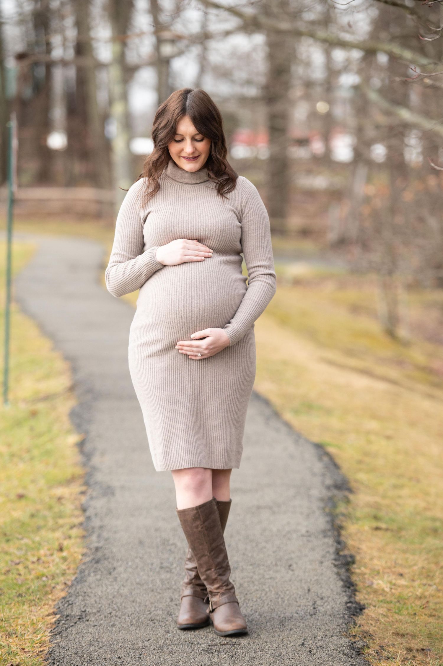 A woman in a grey sweater dress poses for maternity photos on a path during winter.