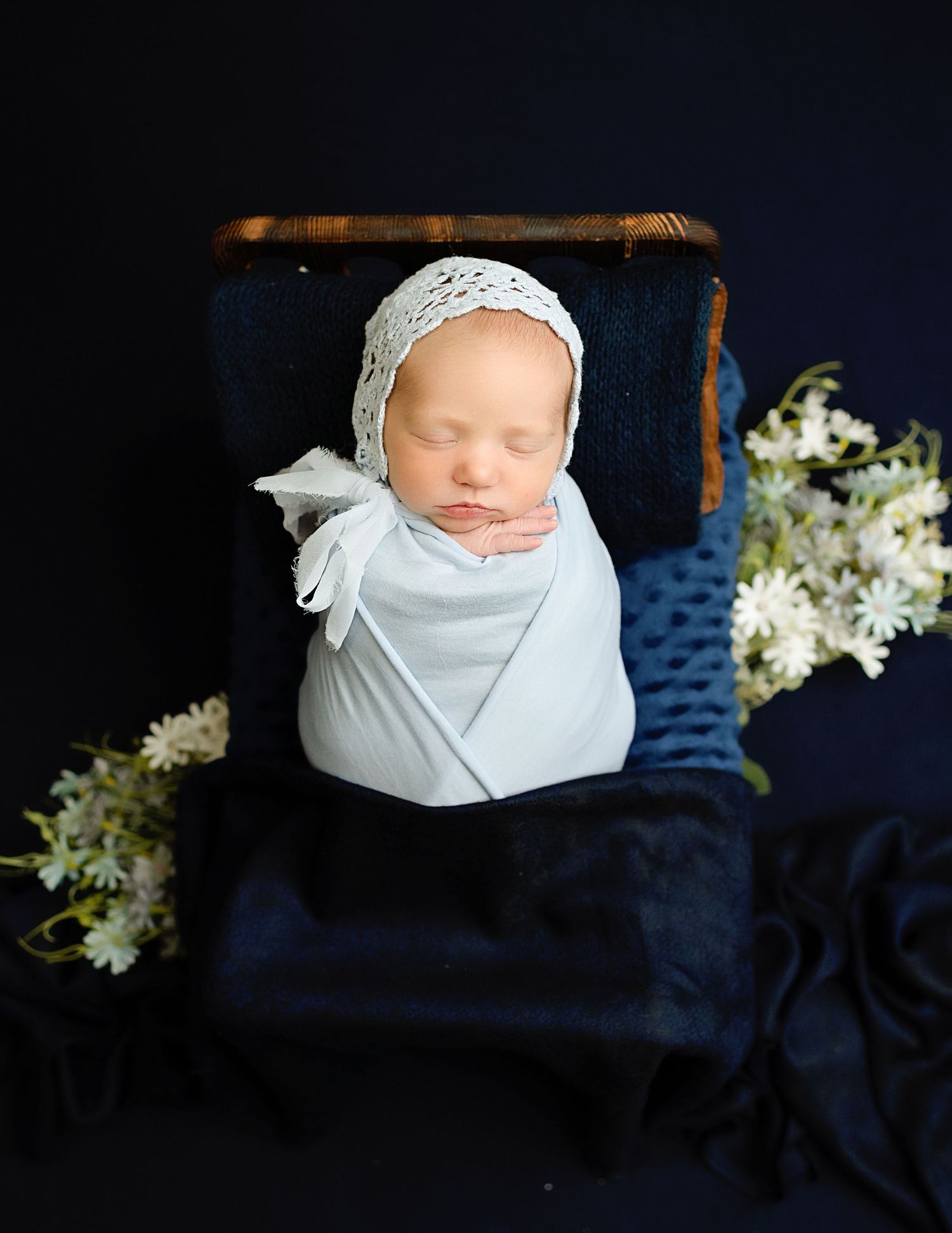 Newborn baby wrapped in white fabric with lace bonnet against dark blue background with white flowers.