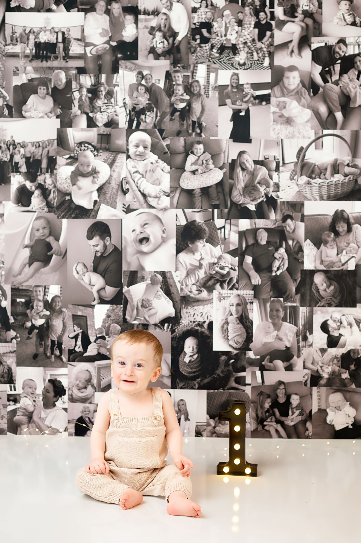 Baby sits in front of photo collage wall with illuminated number 1 during first birthday photoshoot.