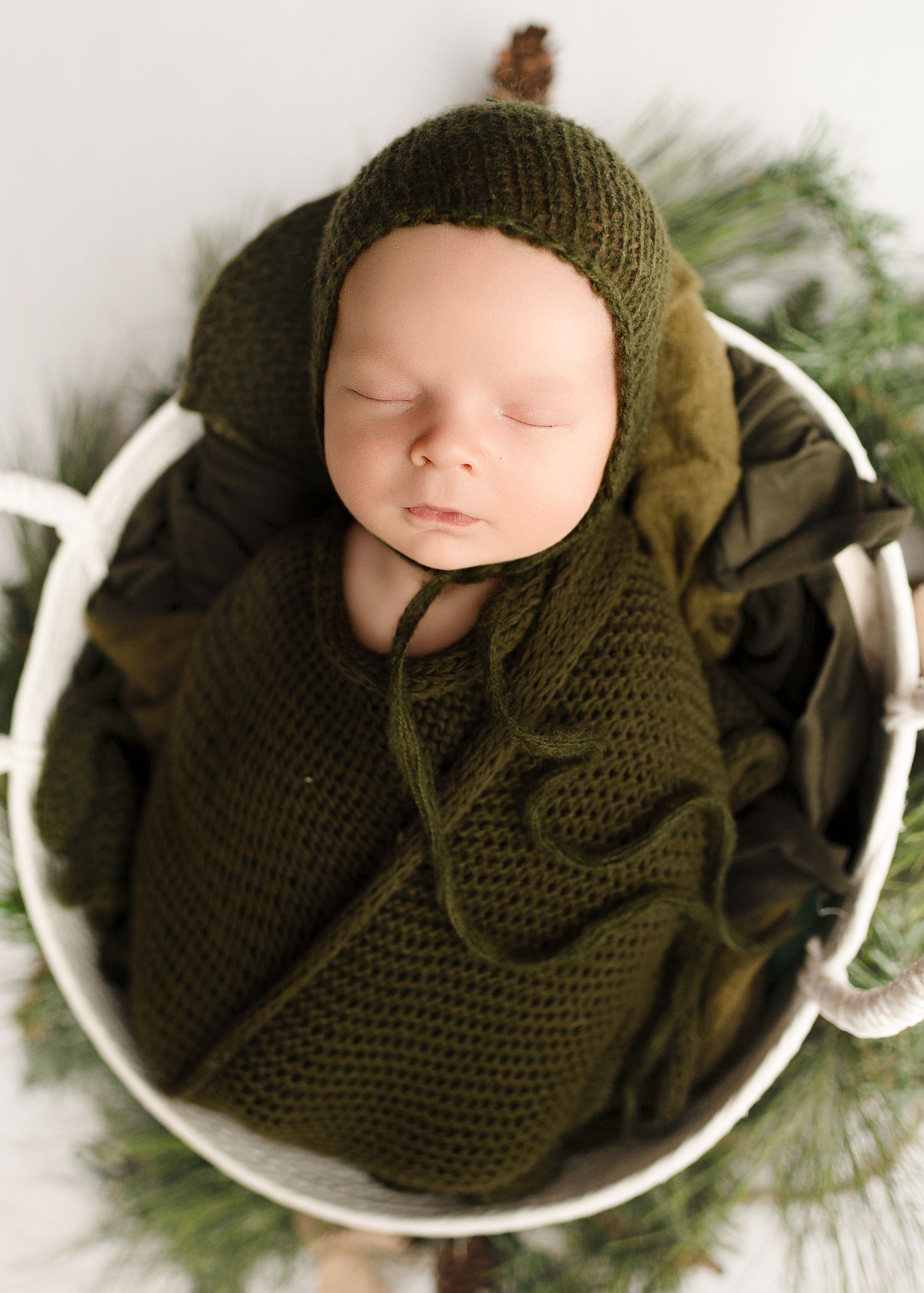 A sleeping newborn swaddled in olive green knit fabric lies peacefully in a white bowl surrounded by evergreen branches.