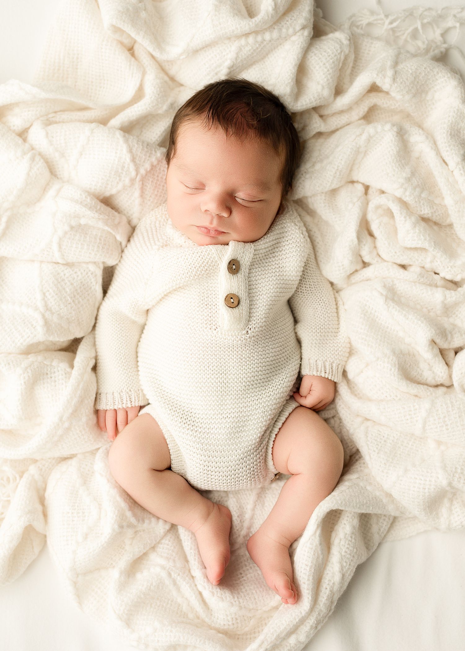 Newborn baby wearing white knit sweater lying on textured cream blanket in studio setting.