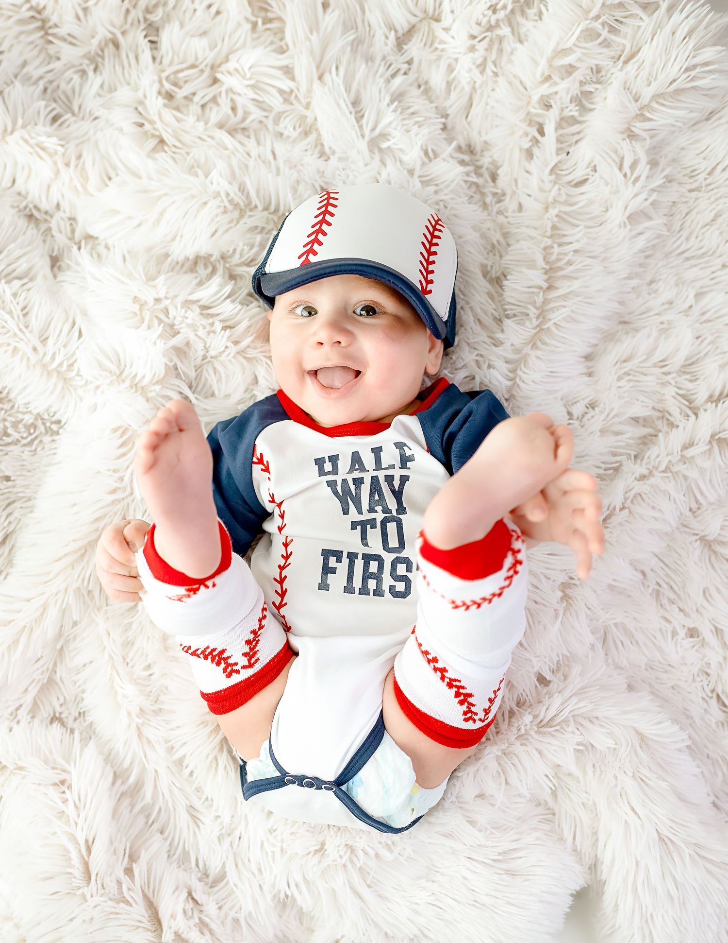 Baby wearing baseball themed onesie lying on white furry rug.