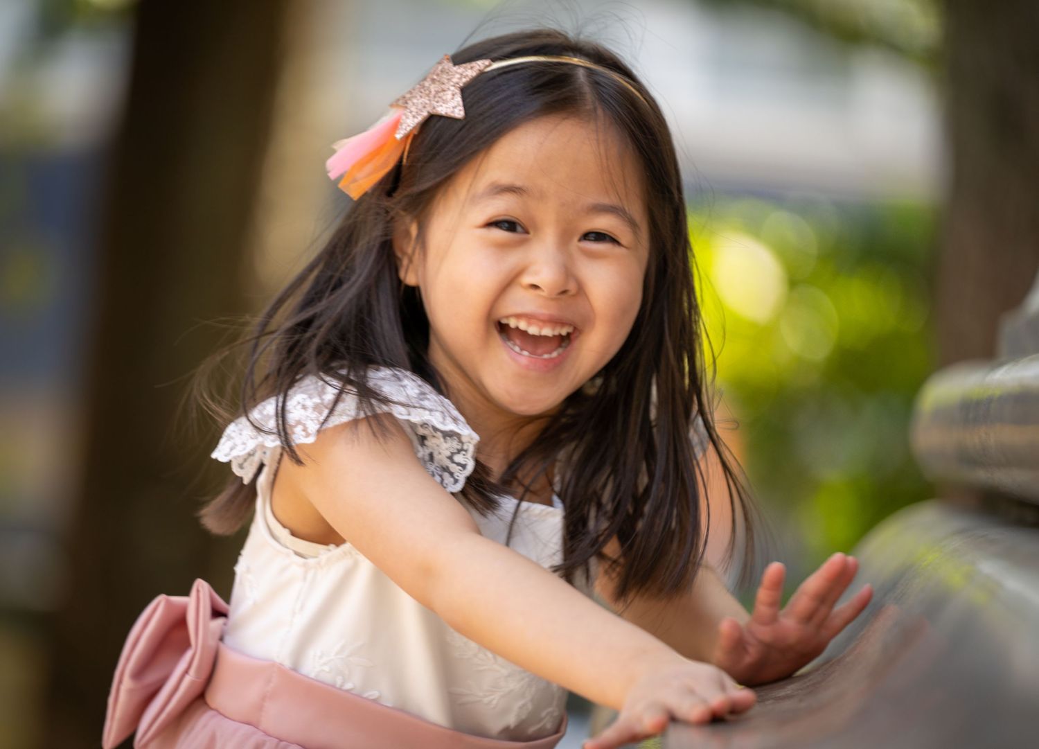 Joyful young Asian girl with pink hair bow laughing and playing outdoors on a sunny day.