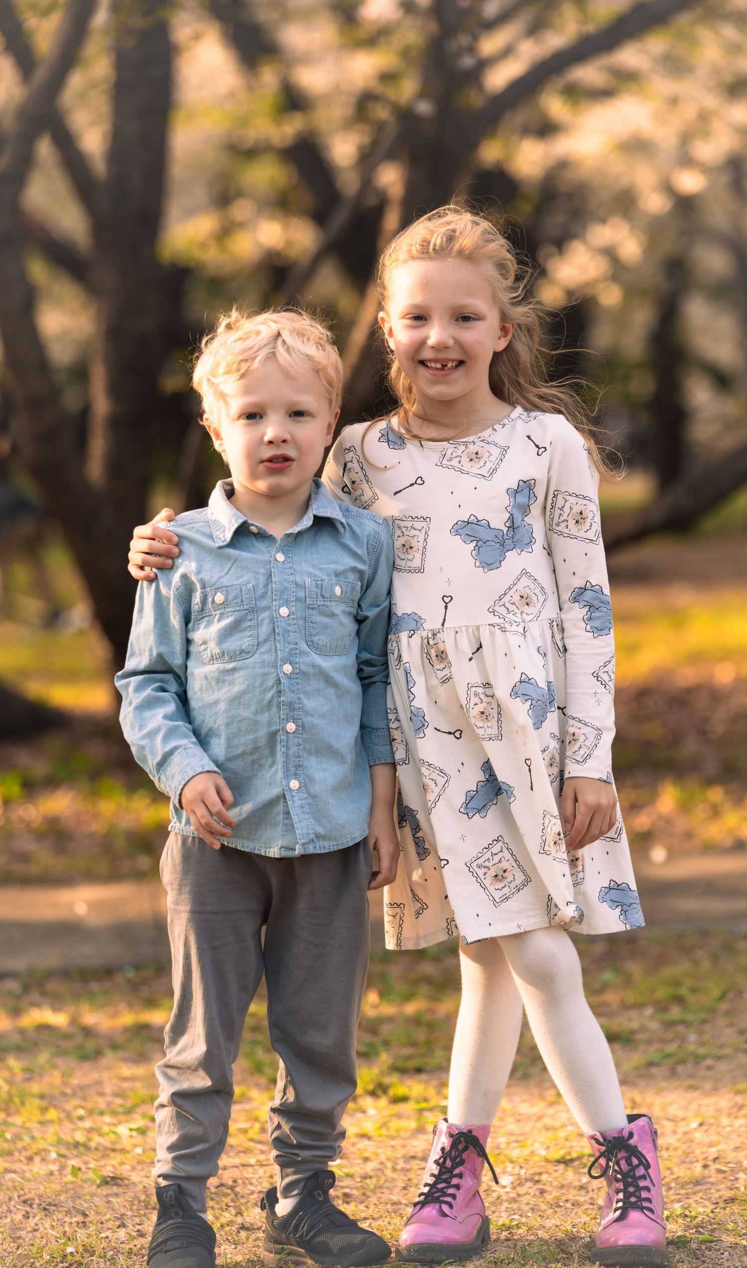 Two children in casual outfits standing together on autumn leaves with purple boots.