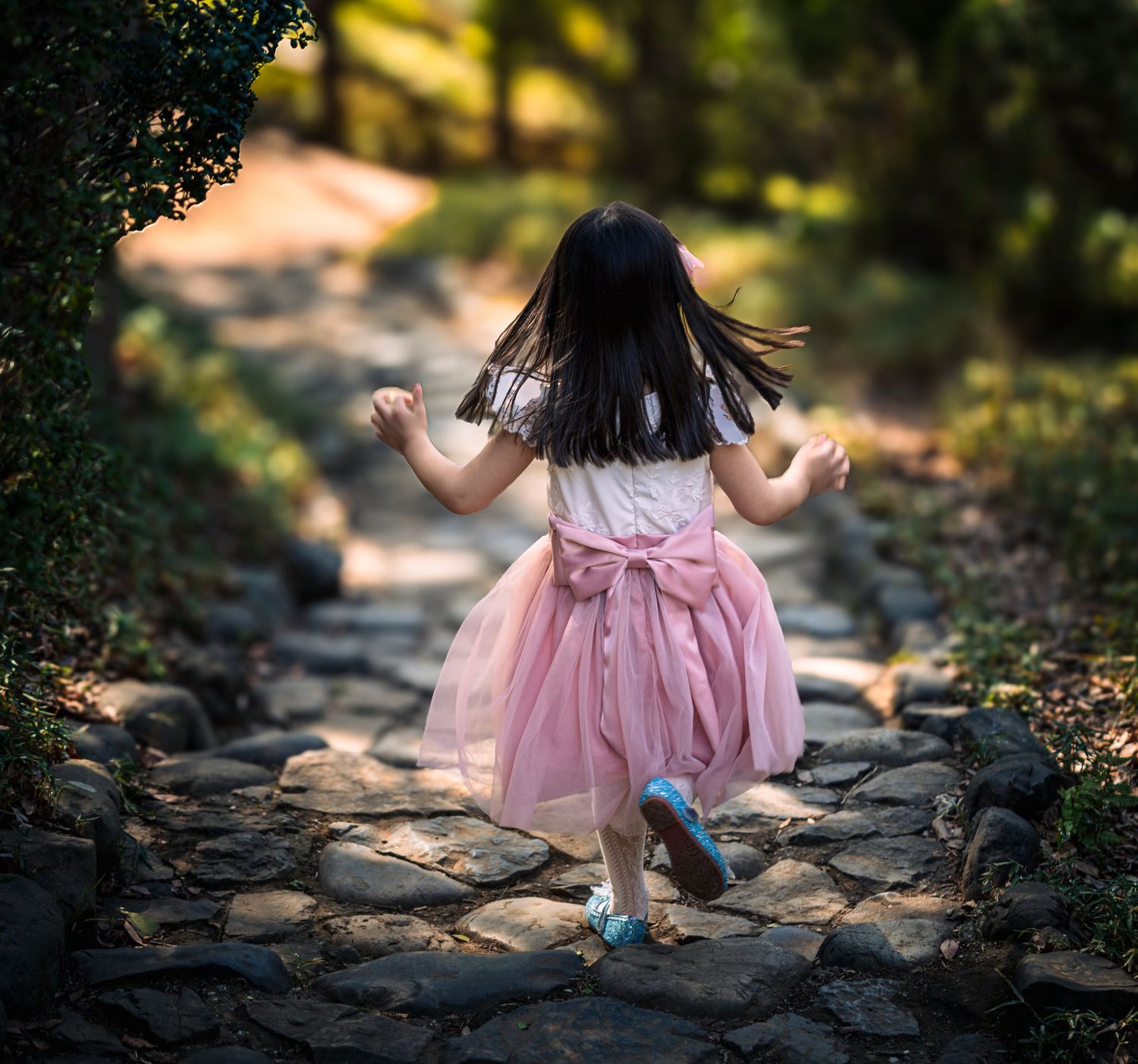 A little girl in a pink tutu dress with a bow runs along a rustic stone path in a lush green garden.