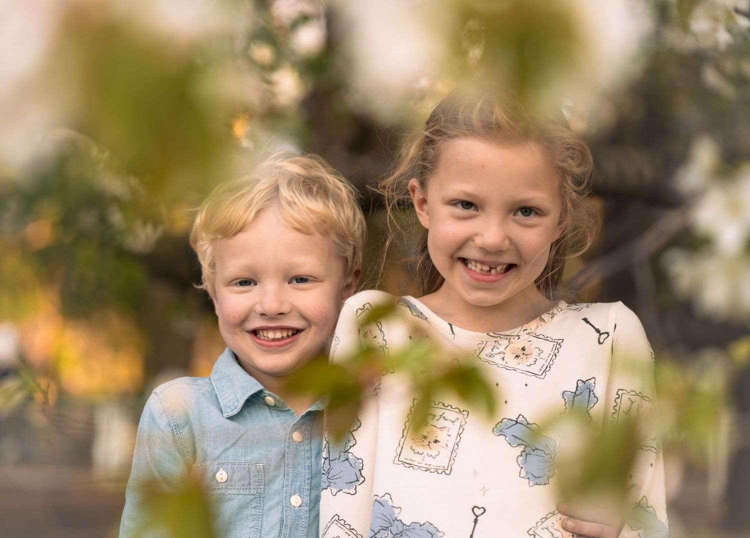 Two children smiling through blurred foreground foliage in a natural outdoor setting.
