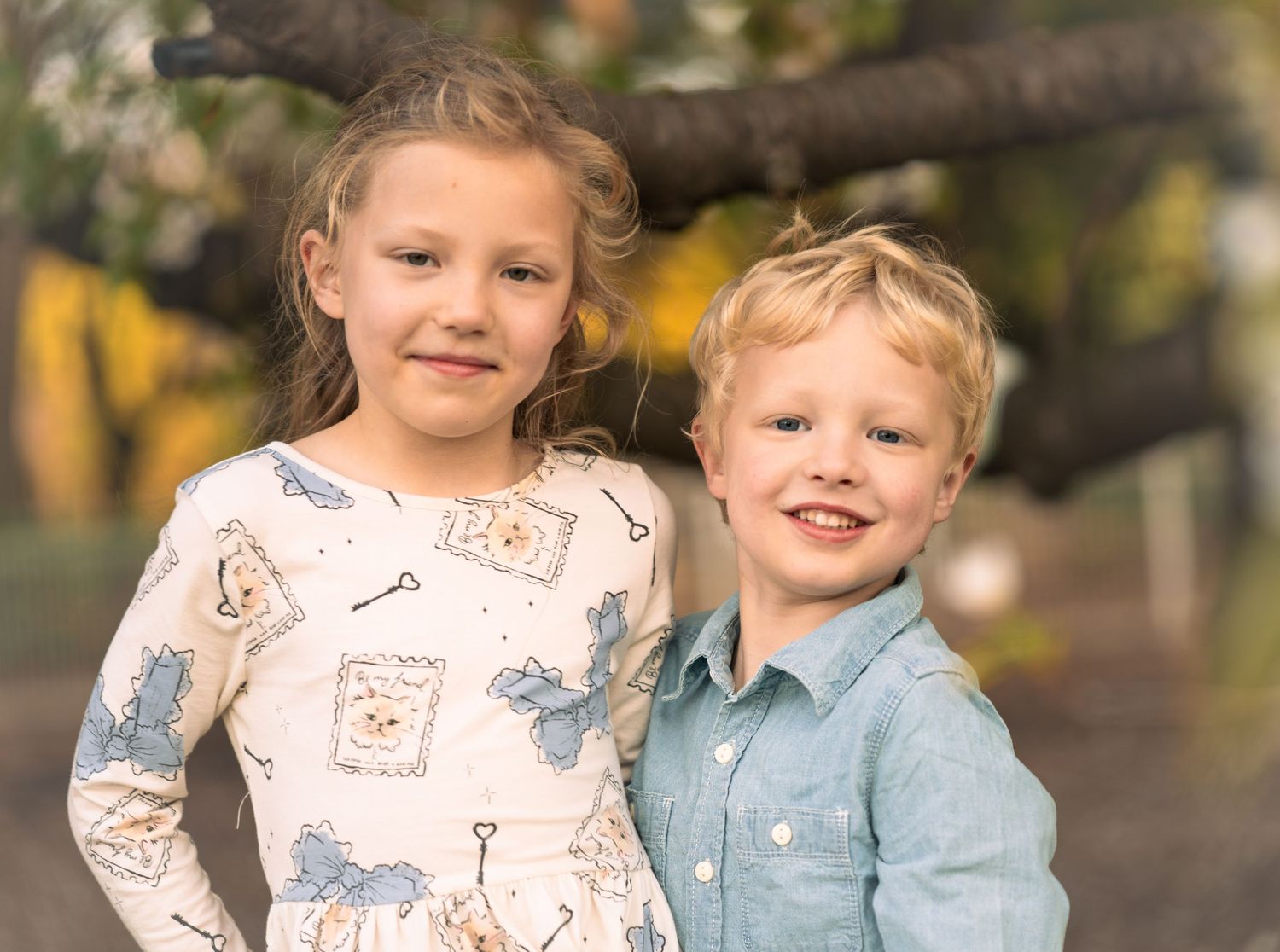 Two young children posing together outdoors with warm autumn colors in the background.