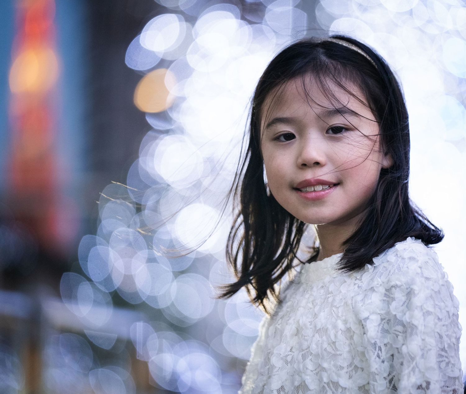 A young Asian girl with dark hair smiles warmly, wearing a white top, with bokeh lights in the background.