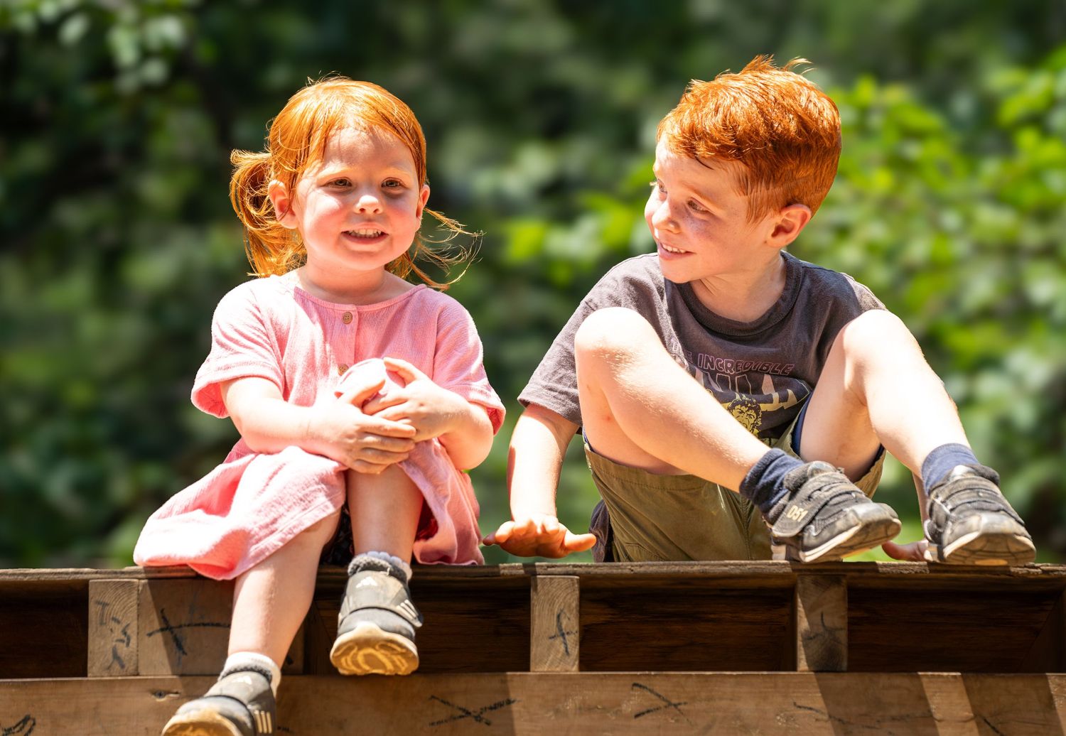Two children sitting on a wooden bench outdoors in sunlight, enjoying a summer day together.