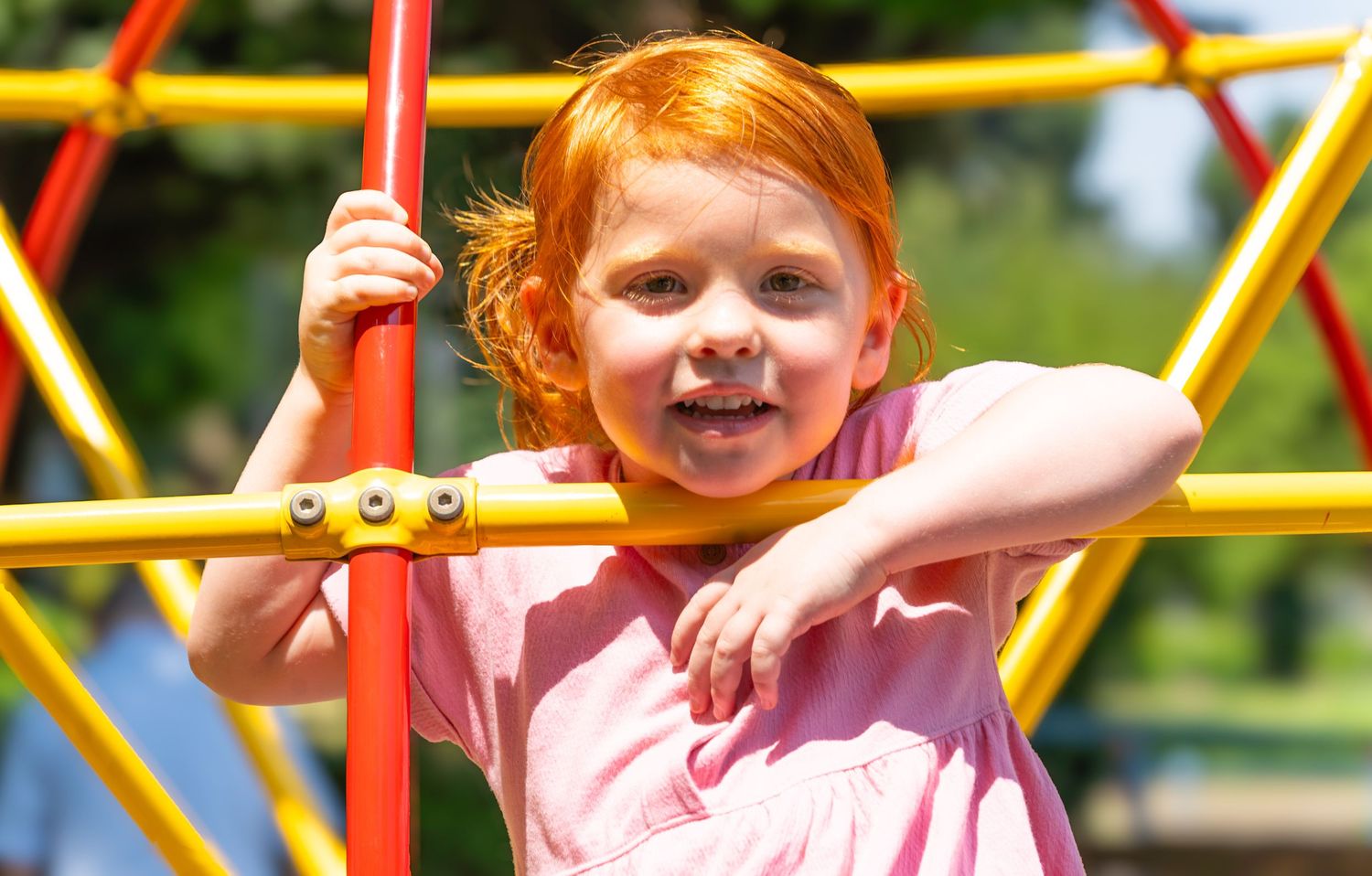 Young child in pink plays joyfully on outdoor climbing equipment at a playground on a sunny day.