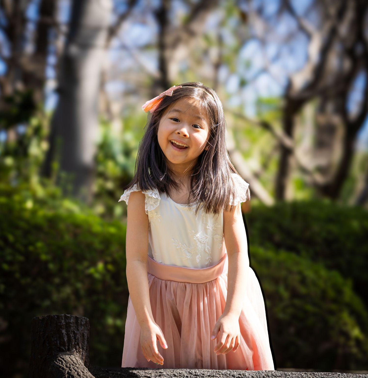 A smiling young Asian girl in a white top and pink tulle skirt poses outdoors in a sunlit garden park.