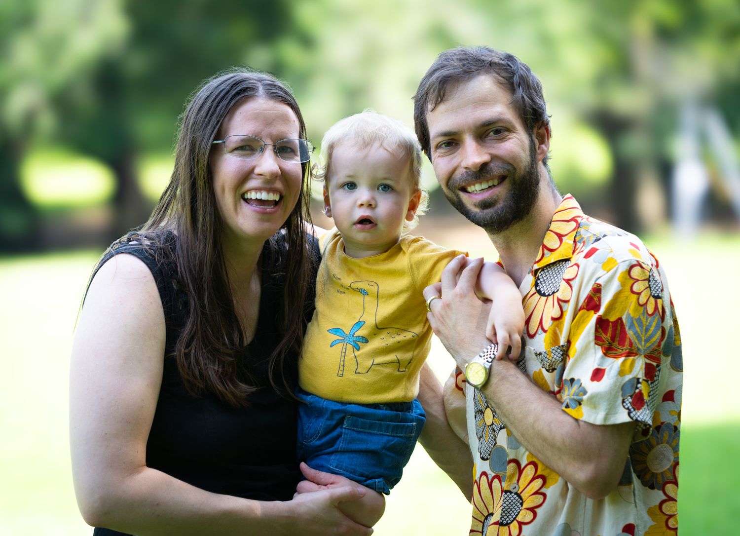 Happy family portrait with mother, father, and toddler smiling outdoors in a lush green park setting.