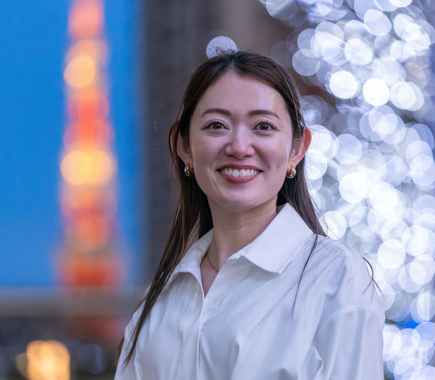 Smiling Asian woman in white shirt poses outdoors at night with Tokyo Tower and bokeh lights behind her.