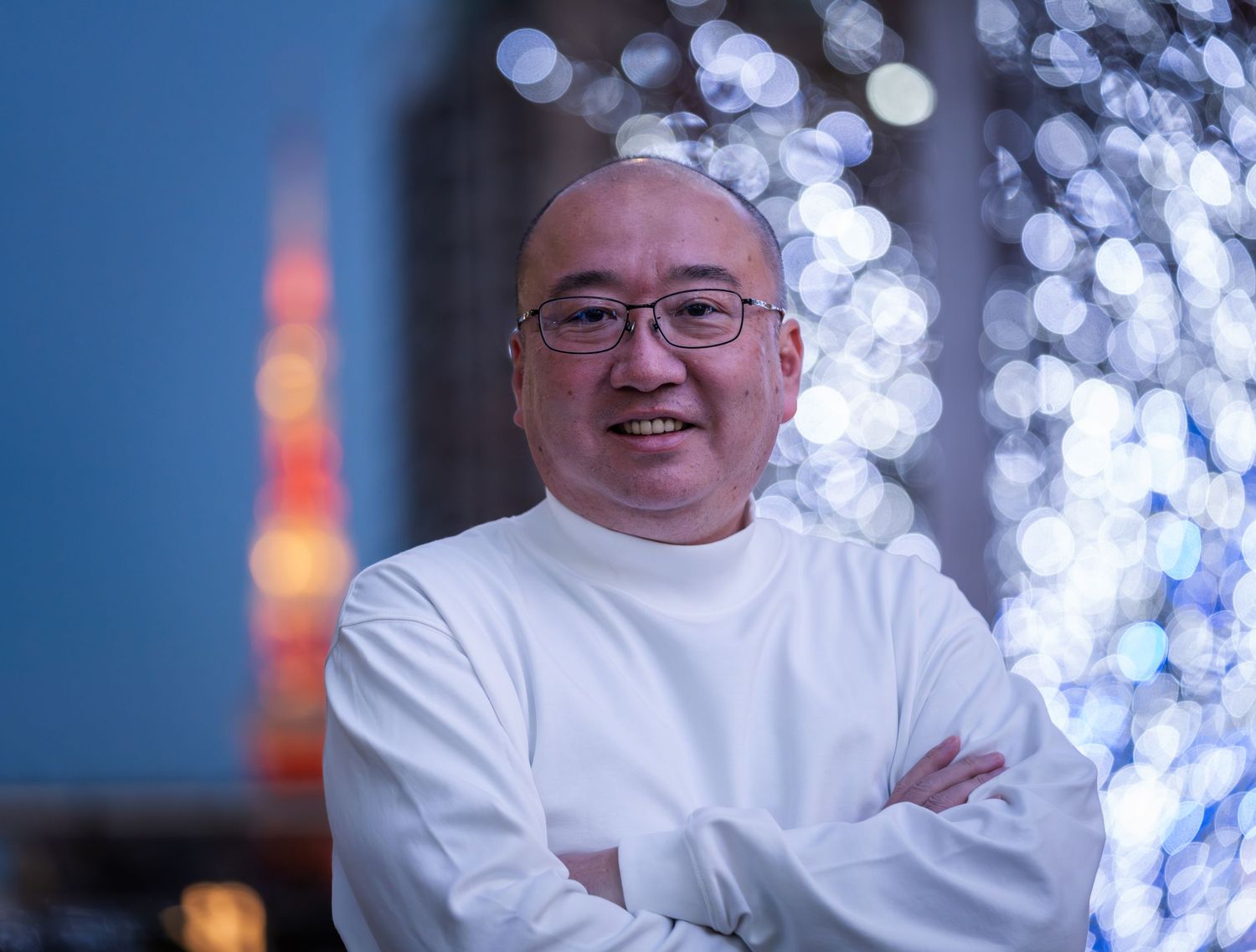 Smiling Asian man in white chef uniform with arms crossed, bokeh lights and Tokyo Tower in background.