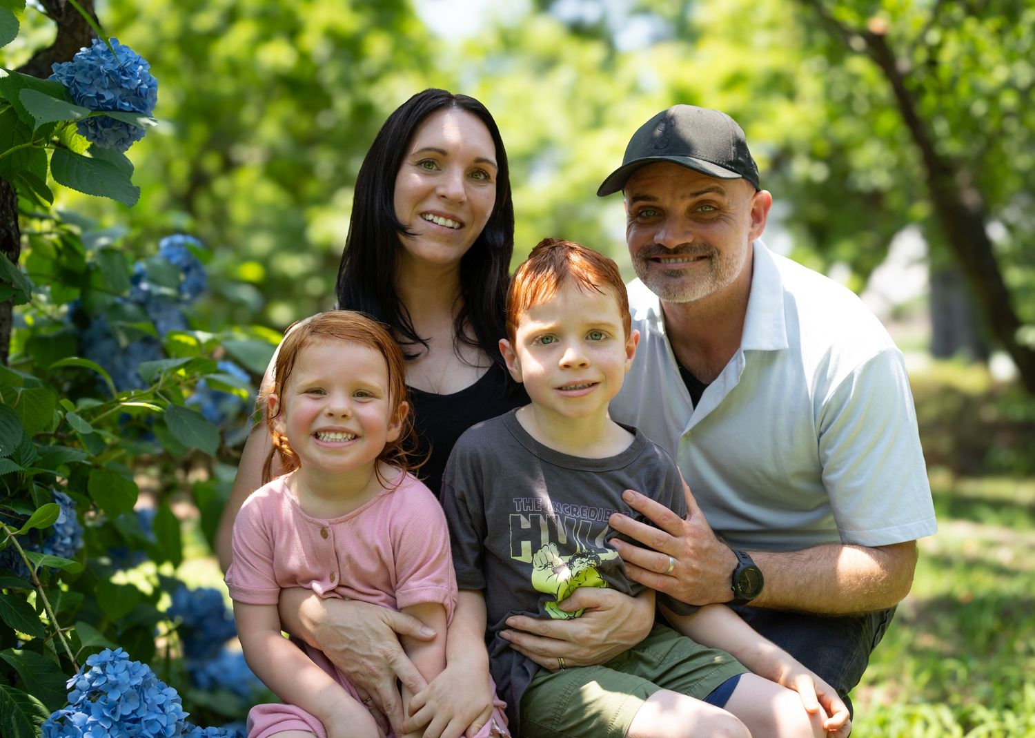 A happy family sitting outdoors in a lush garden with blooming hydrangea flowers on a sunny summer day.