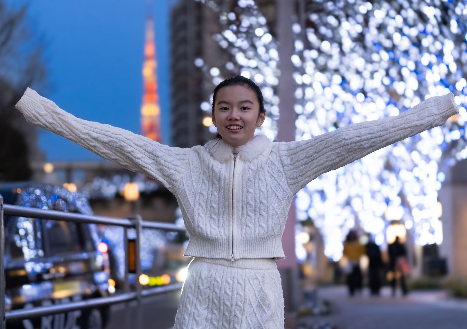 Young woman in white cable-knit sweater poses with arms outstretched among festive lights near Tokyo Tower at dusk.