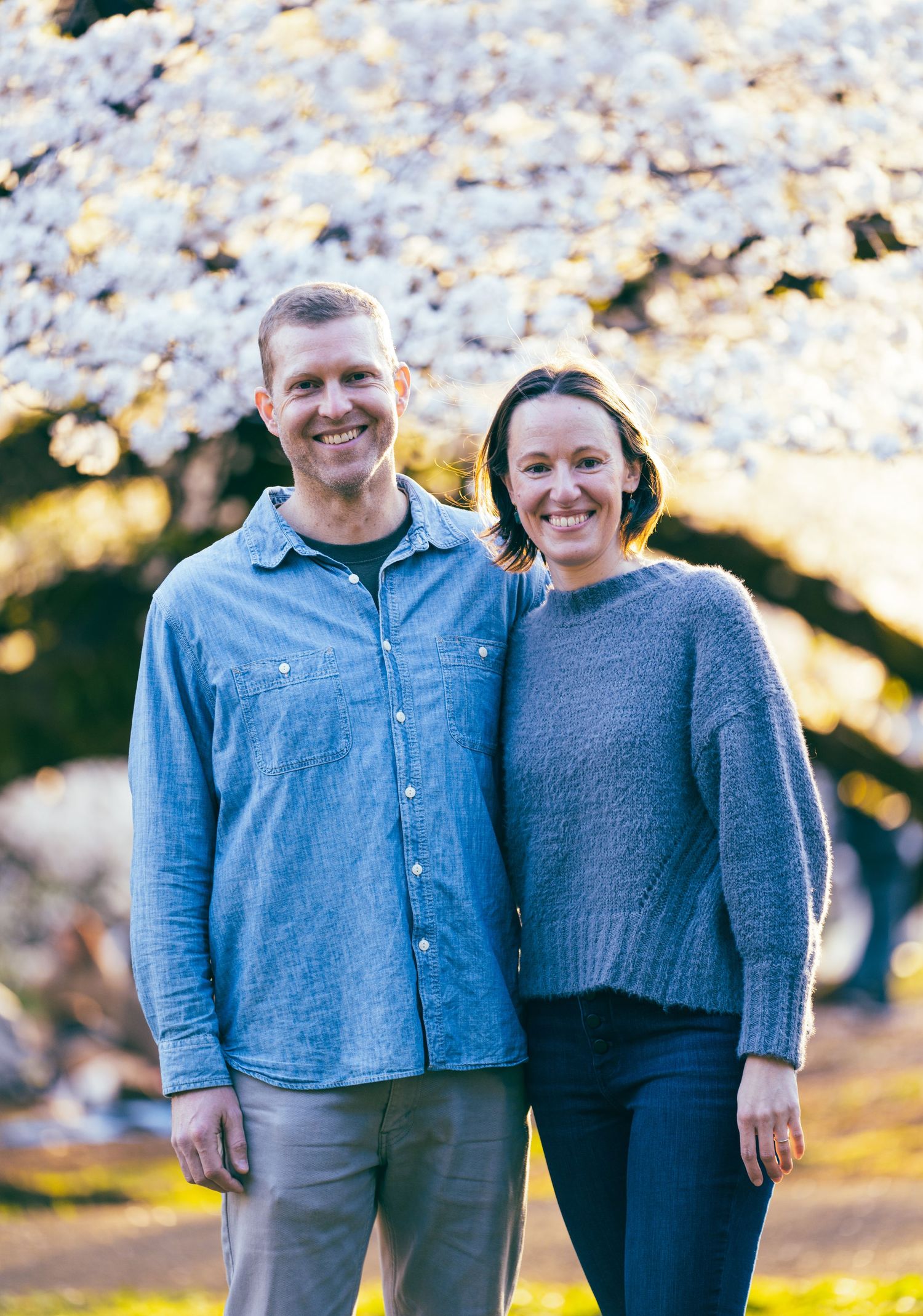 A couple smiles together under blooming cherry blossoms on a sunny day.