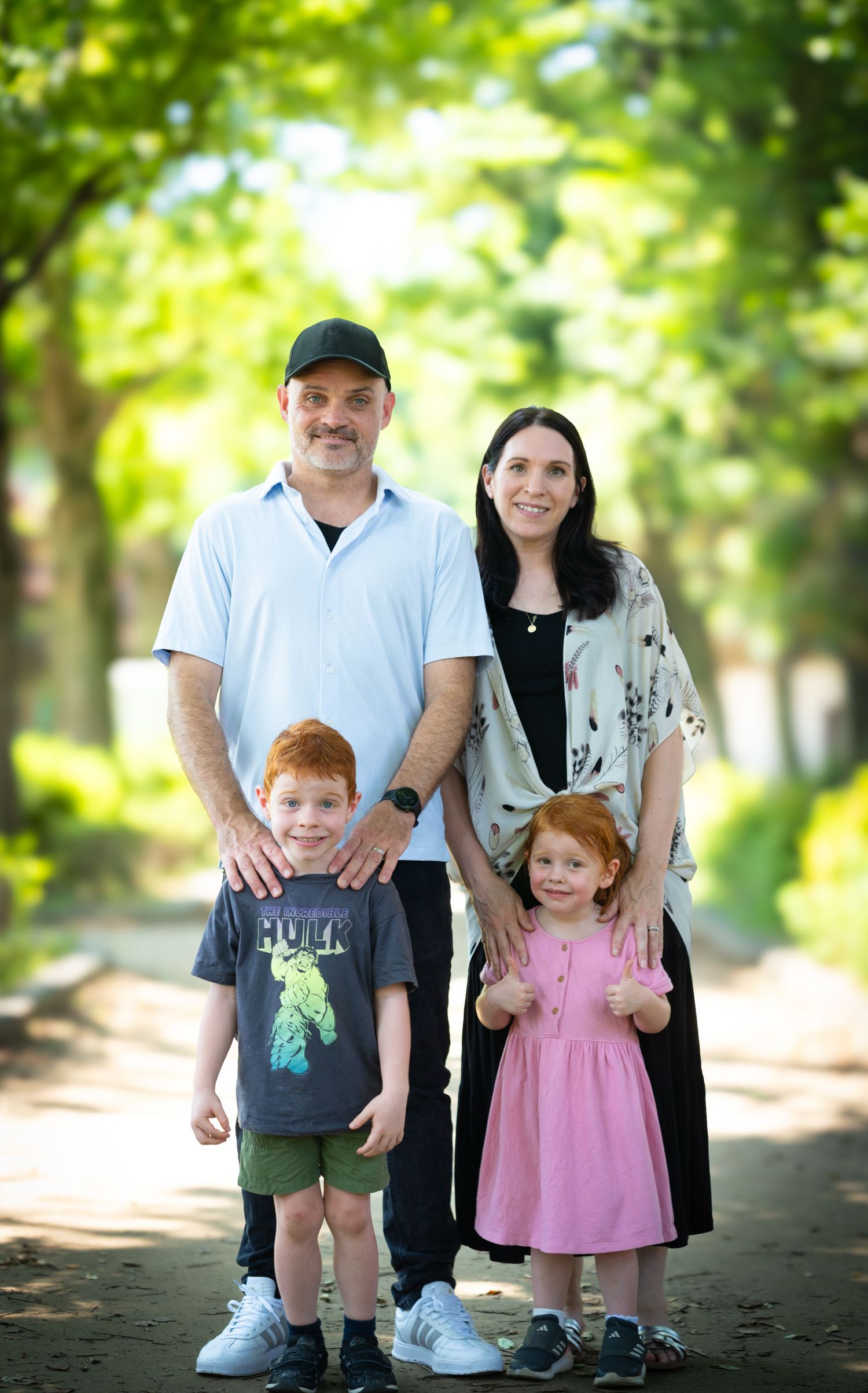 A family stands together on a tree-lined path with greenery and dappled sunlight in the background.